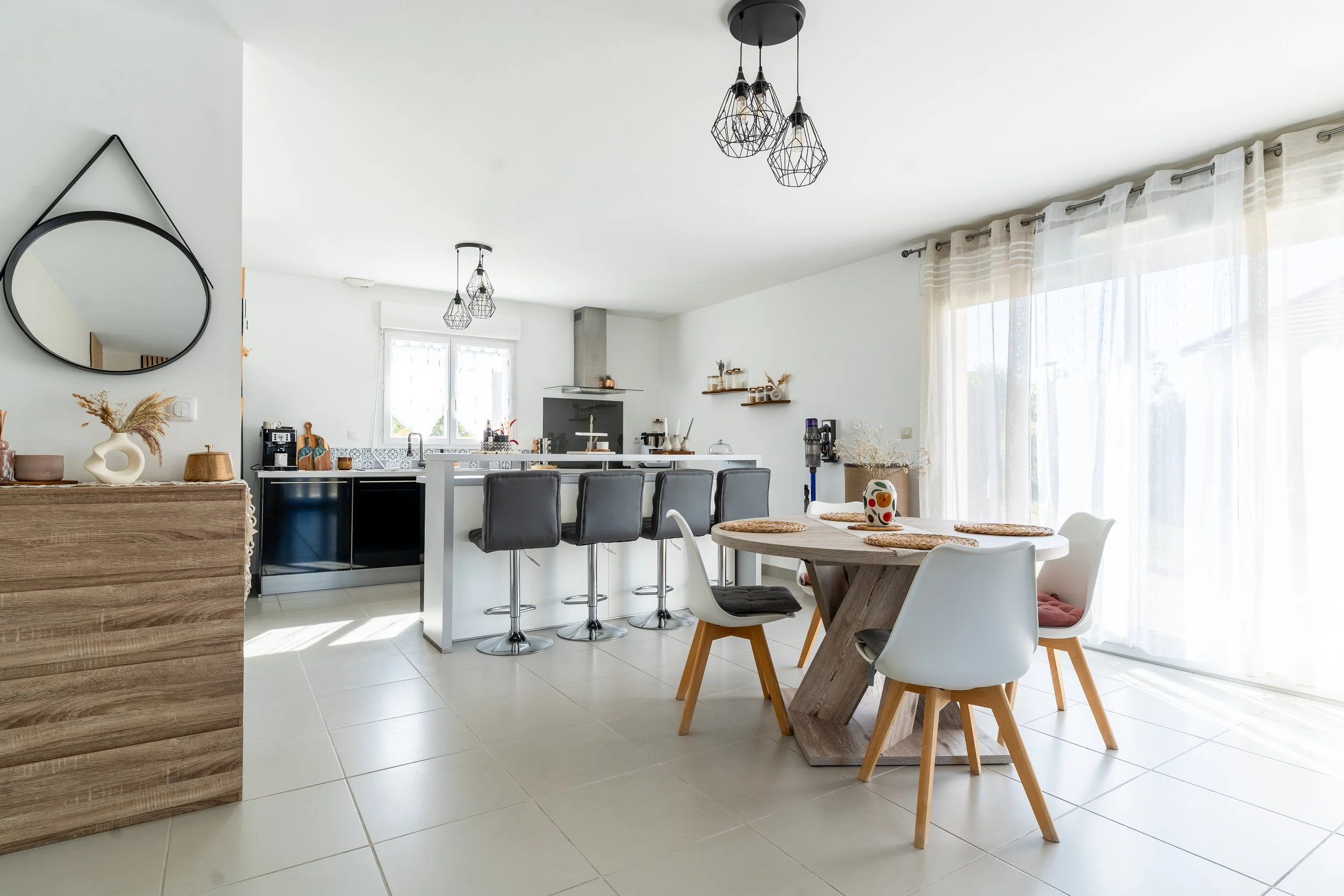 Une cuisine ouverte et lumineuse avec un coin repas, des chaises modernes, un plan de travail avec des tabourets, et des décorations minimalistes. Grandes fenêtres avec rideaux translucides, éclairage suspendu en forme de cages en métal.