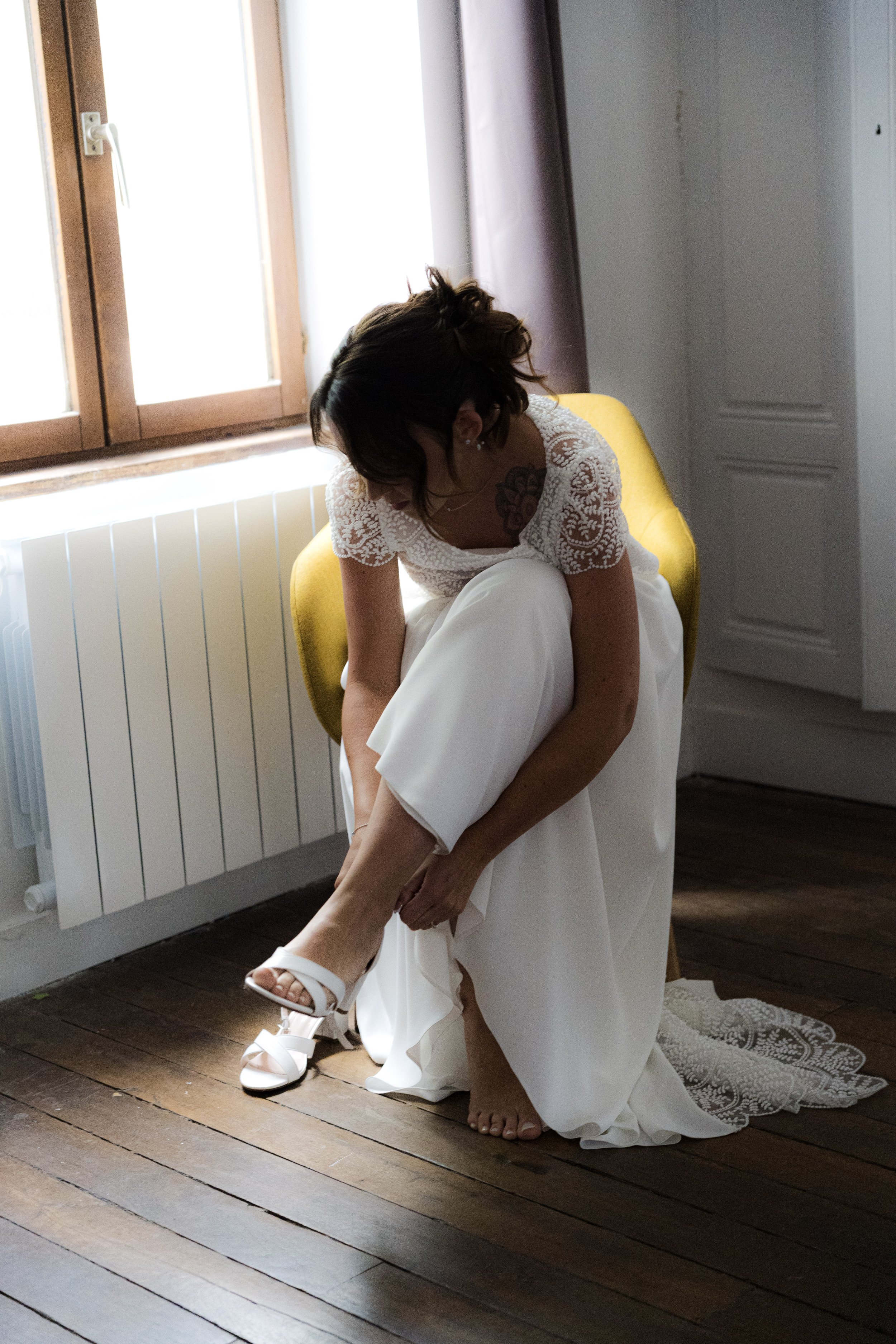 Une femme en robe blanche ajuste sa chaussure sur un meuble en bois, dans une pièce lumineuse avec une fenêtre et un radiateur.