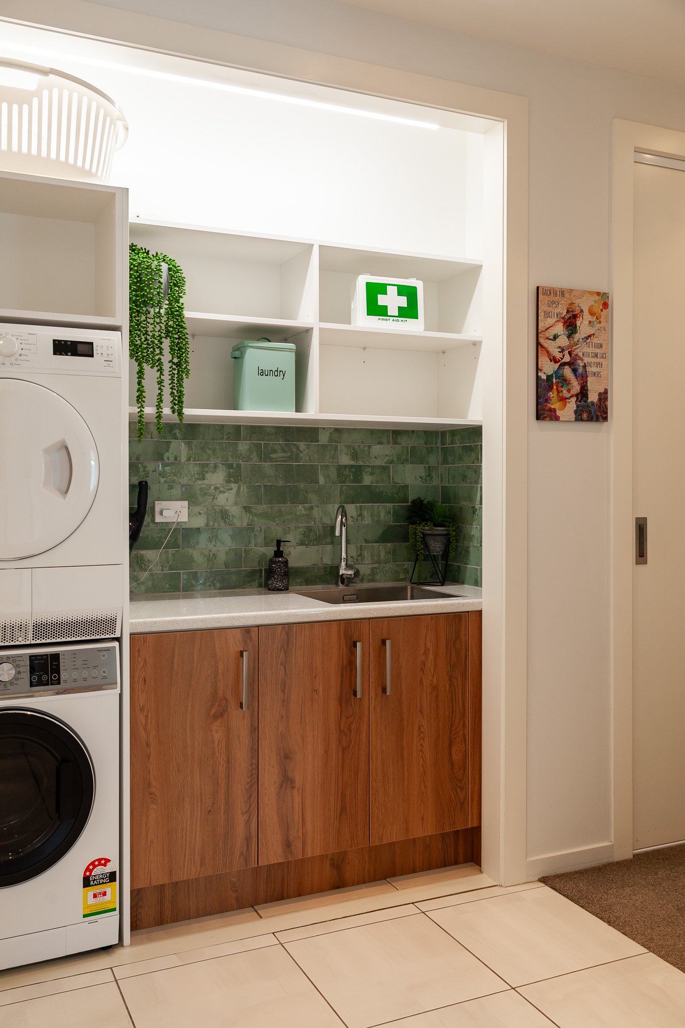A laundry room with a washer and dryer, a kitchen sink with a green backsplash, wooden cabinets, and shelves holding a green plant, a first aid kit, and laundry supplies.