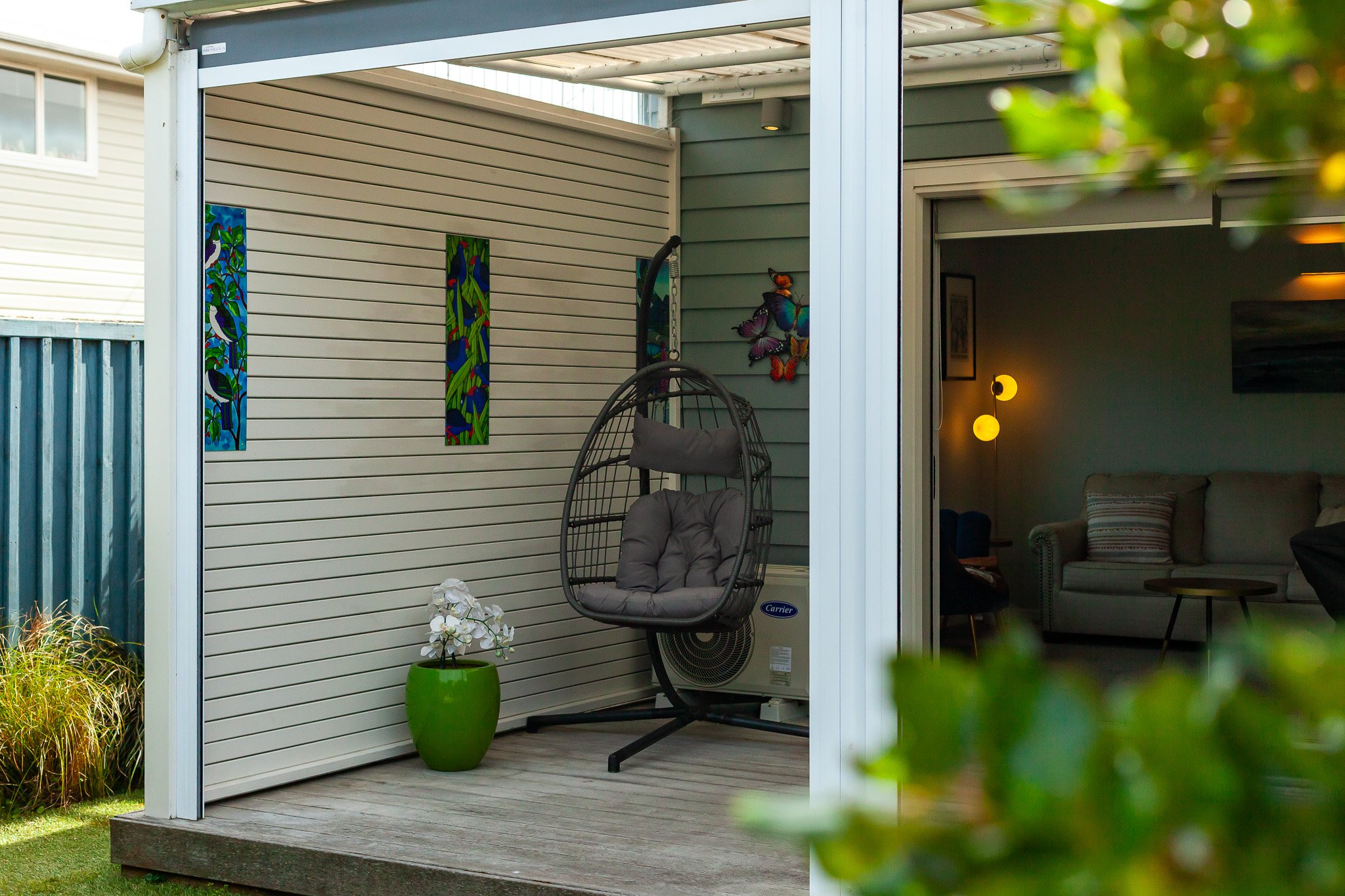 An outdoor patio area with a hanging egg chair with cushions, a green plant in a green pot, and colorful wall art on a wall, partially covered by a roof. Inside, a living room with a beige sofa, striped pillows, and a floor lamp with yellow globe lights is visible through an open sliding door.