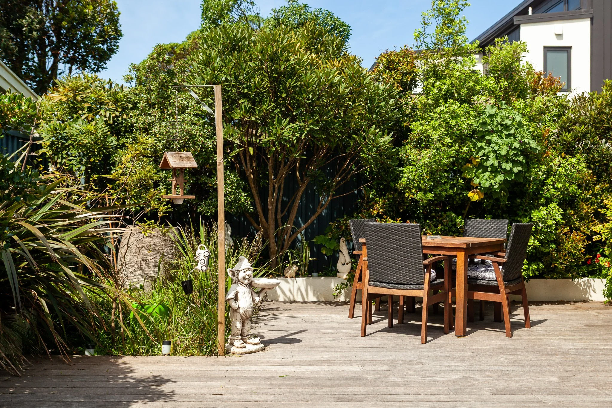 A wooden outdoor patio with a table and four chairs surrounded by lush green bushes and trees, a garden gnome statue, birdhouse on a tall pole, and various decorative plants.