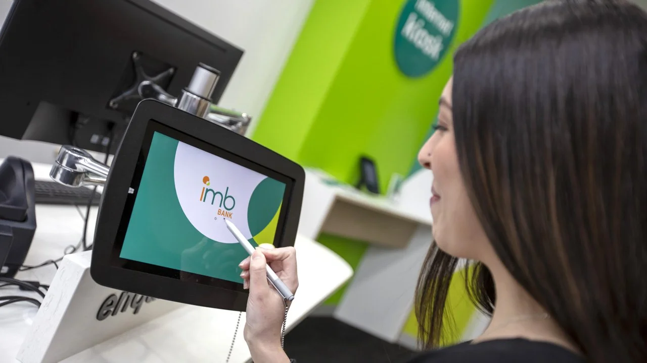 A woman is holding a stylus and interacting with a touchscreen monitor displaying the IMB Bank logo at a bank counter.