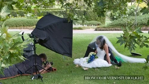 A wedding photo shoot in progress, featuring a photographer on the ground capturing a bride in a white dress and veil. A large reflector and studio light are set up nearby, in an outdoor garden setting.