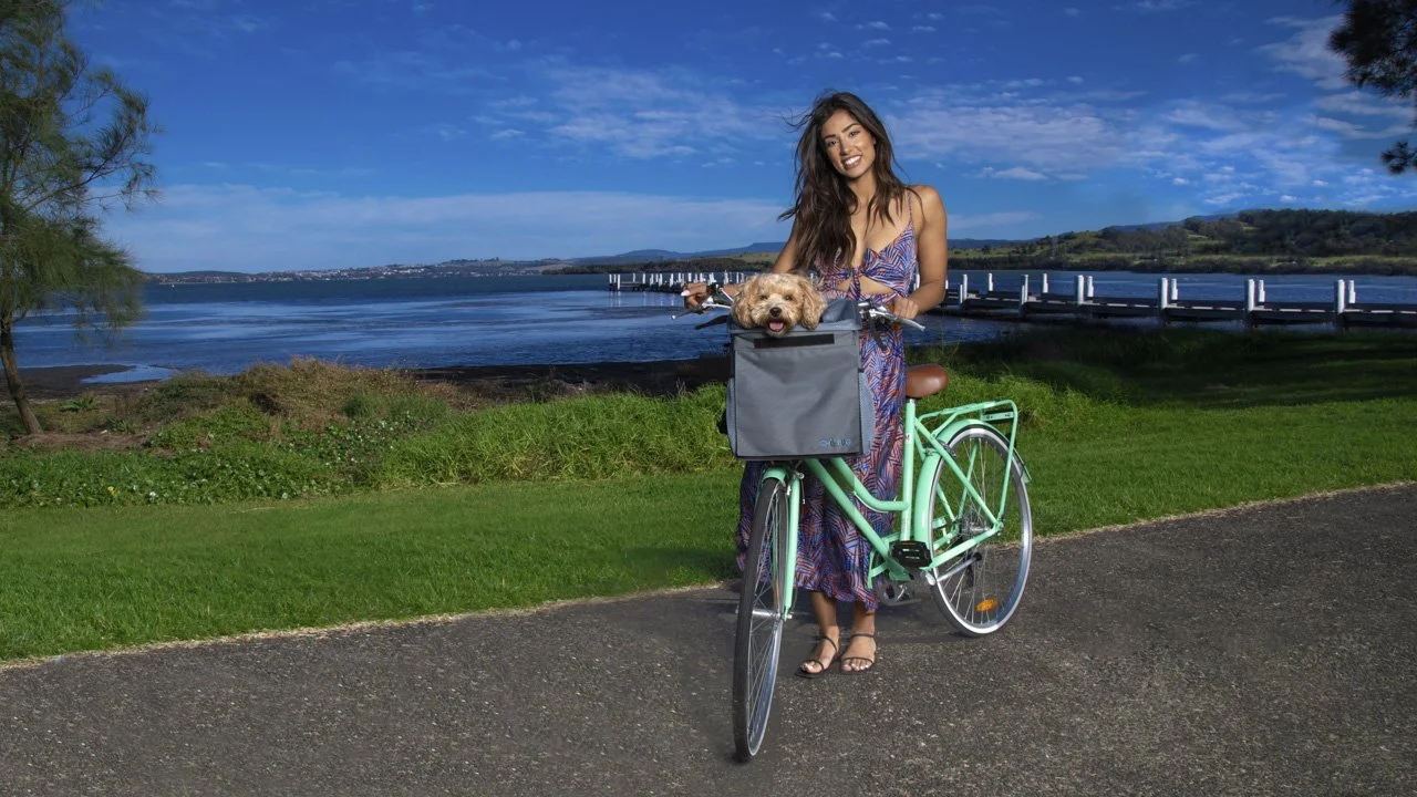 Smiling woman standing with mint green bicycle and Chillbug pet carrier holding a Moodle dog, with a scenic lake and boardwalk in the background – lifestyle product image captured by Cole Studios.