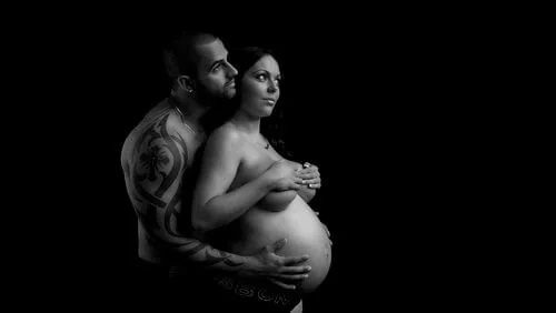 Black and white photo of a pregnant couple embracing; the man has tattoos, and the woman is topless, covering her chest.