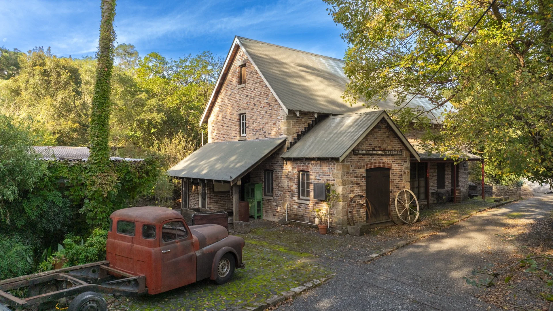 A rustic brick building with a metal roof, surrounded by trees with autumn leaves, and an old rusted truck parked in front.