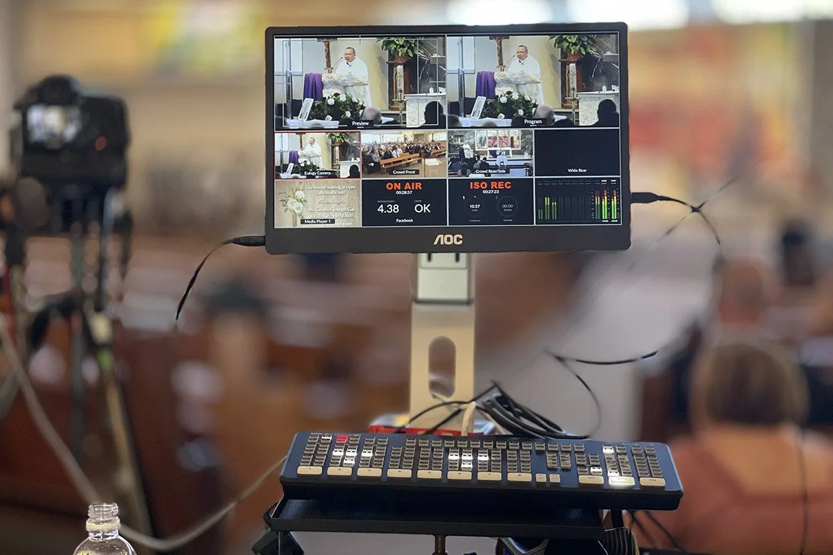 A live video production monitor showing multiple camera angles of a speaker at a lectern in a church or conference setting, with a keyboard and other equipment in the foreground.