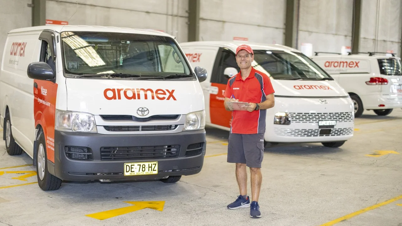 A man in a red Aramex uniform holding a clipboard stands inside a warehouse with white delivery vans that have Aramex branding and logos, parked in marked parking spots.