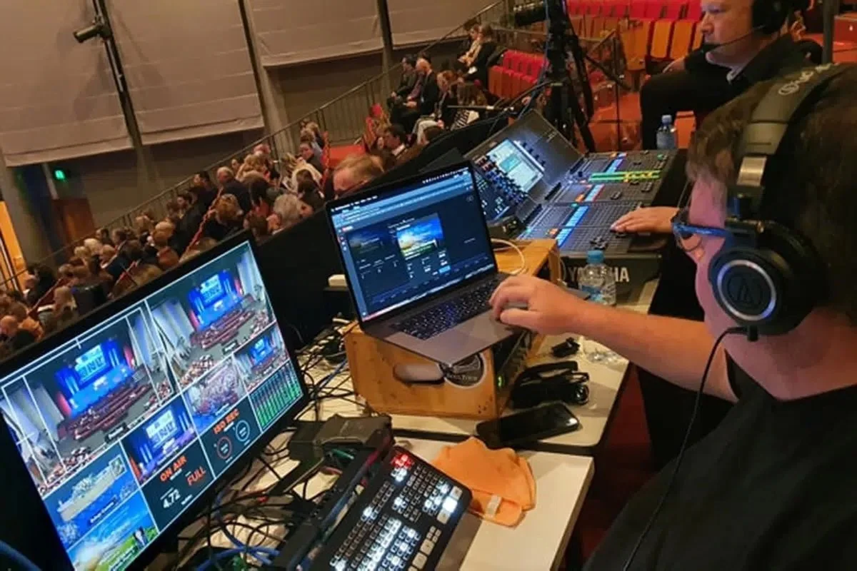 A live event control room with multiple monitors, laptops, and audio equipment, overlooking an audience seated in tiers, with a person working on a laptop wearing large headphones.