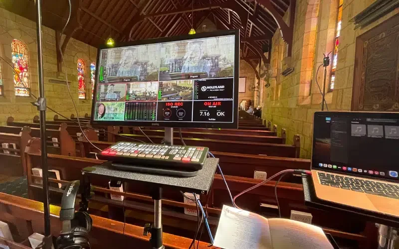 Inside a church with stained glass windows, an electronic control setup with monitors, a keyboard, and a laptop on a pew, likely for recording or streaming services.