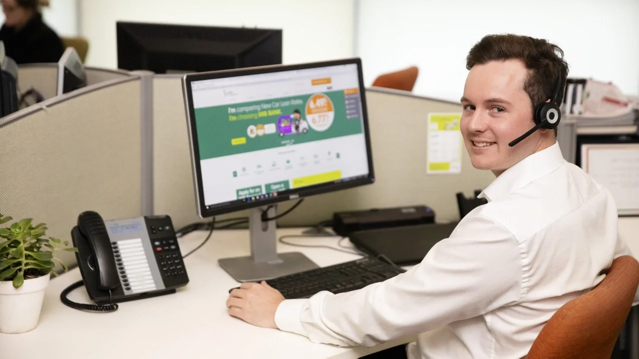 A man wearing a white shirt and a headset sits at a cubicle with a computer monitor, telephone, and a small potted plant, smiling at the camera in an office setting.