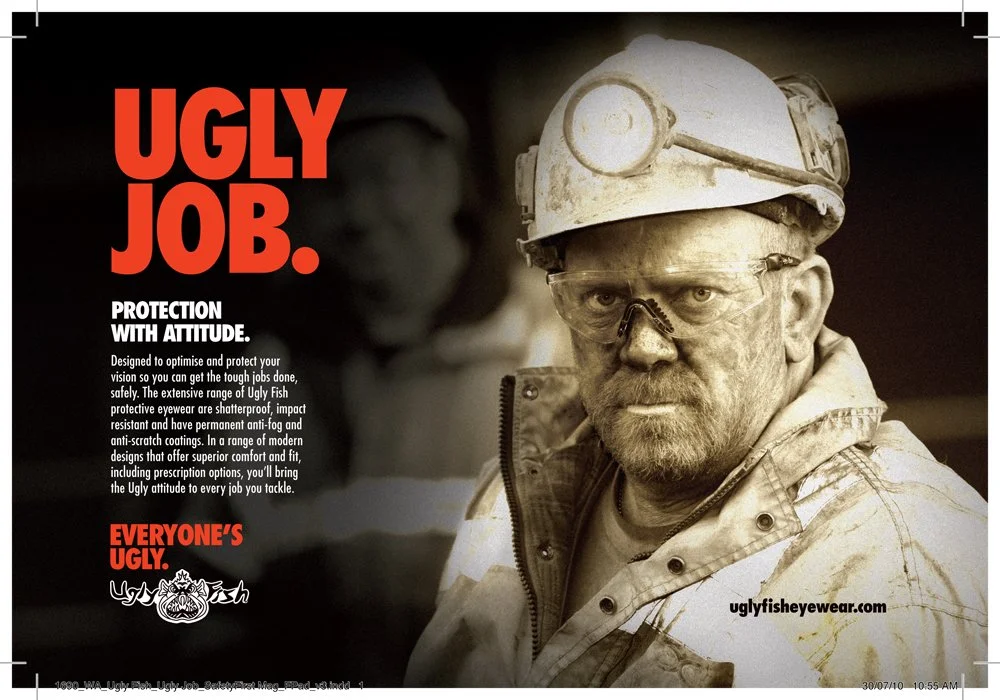 A man in work gear with safety glasses and a helmet, looking serious, advertisement with large red text saying 'UGLY JOB.' for Ugly Fish protective eyewear.