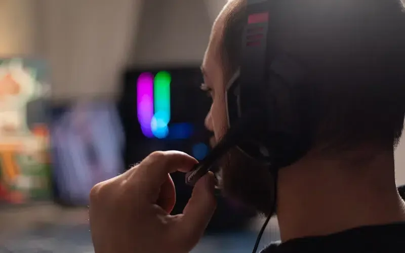A person wearing a gaming headset, seen in profile, adjusting the headset, sitting in front of a colorful monitor.