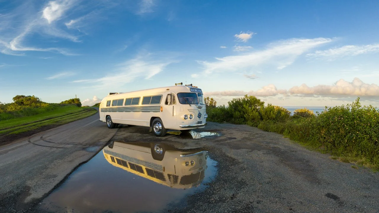 An old white school bus parked next to a puddle on a scenic roadside with green bushes, a blue sky with scattered clouds, and the ocean in the distance.