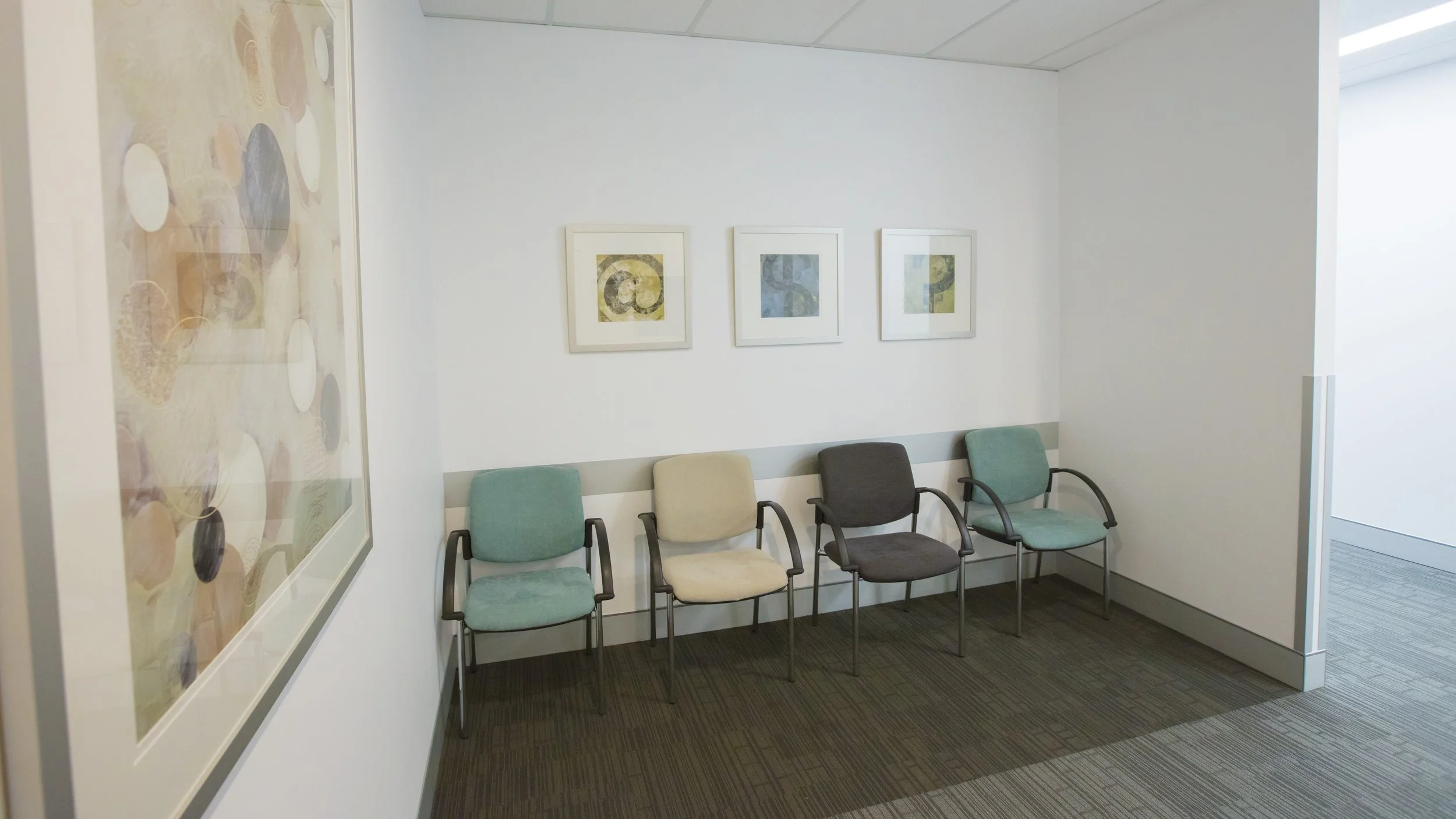 Hospital waiting room with five empty chairs, artwork on the walls, and a carpeted floor.