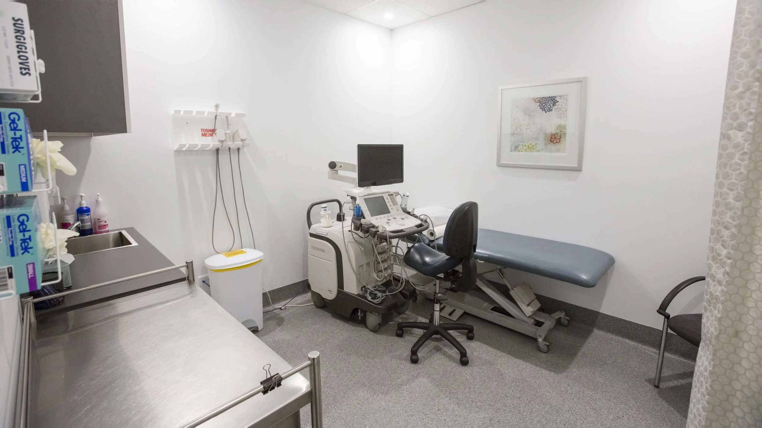 A blank hospital examination room with a medical stool, examination bed, ultrasound machine, sink, and medical supplies.
