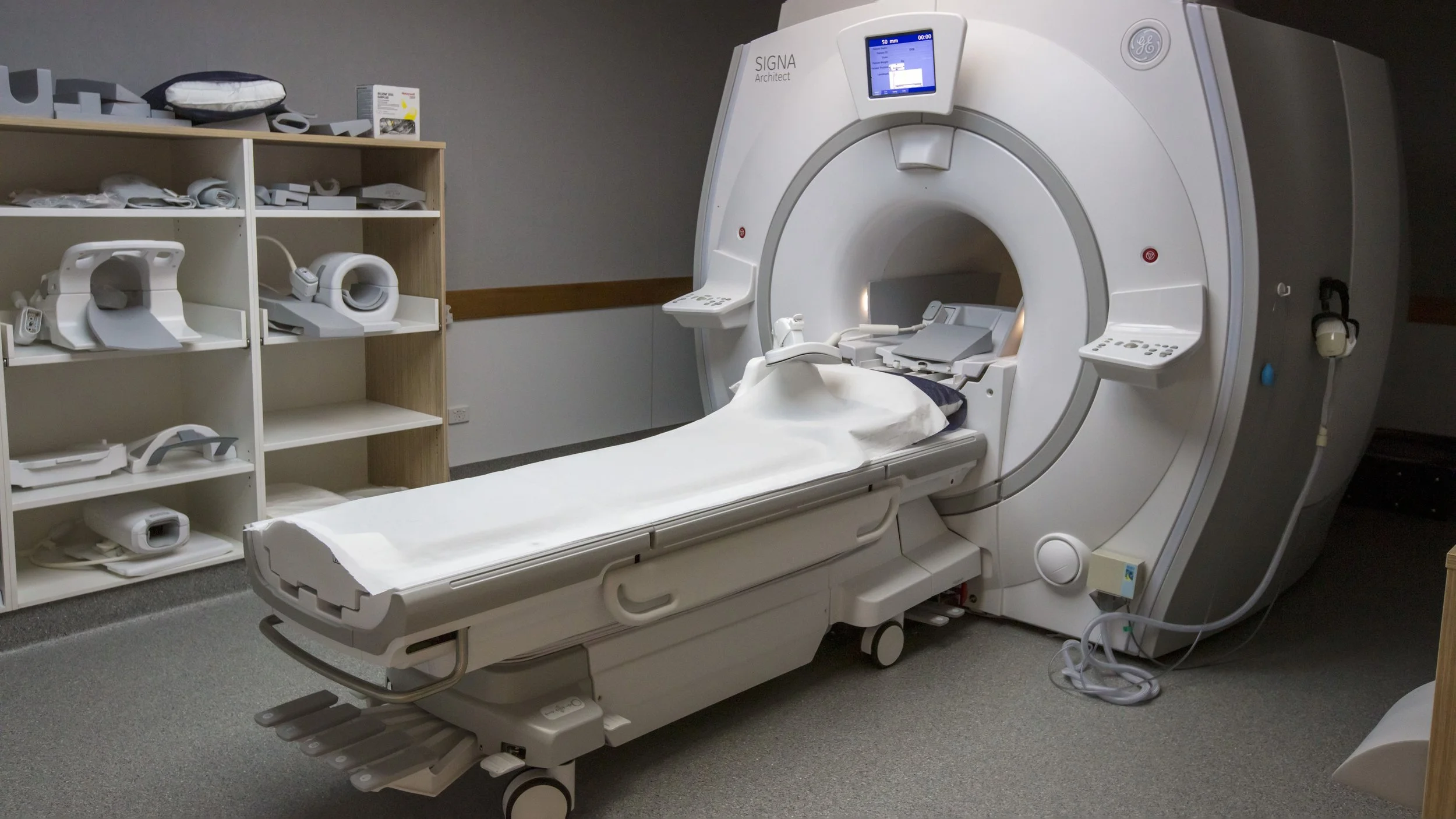 An MRI machine in a medical imaging room with a patient bed and a shelf holding various MRI parts.