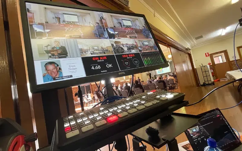A video production control panel with a monitor showing multiple camera feeds, a keyboard, and other electronic equipment in a room with wooden walls and large windows.