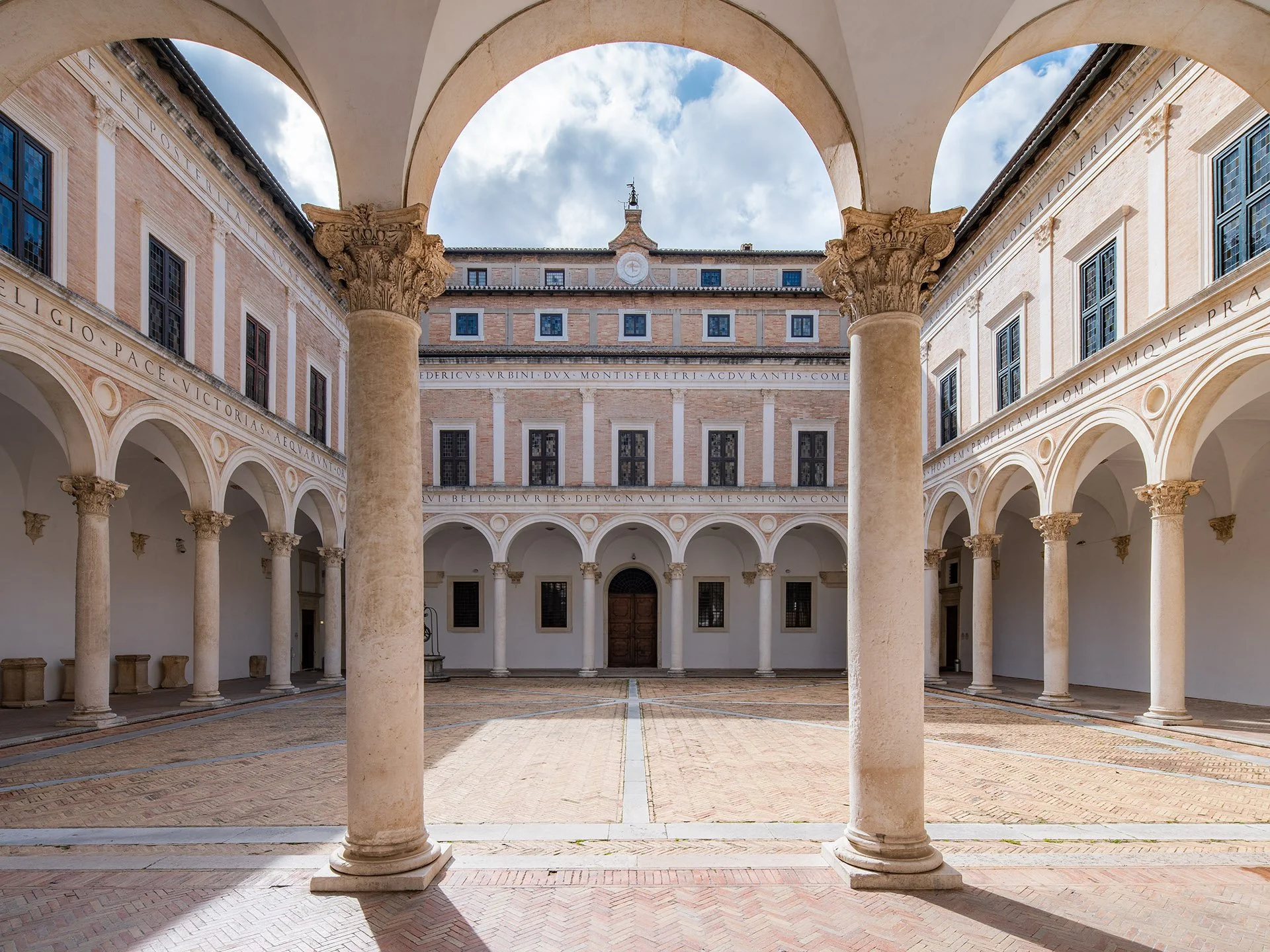 A historic courtyard with columns and arches, surrounded by classical buildings with Latin inscription engravings, under a partly cloudy sky.