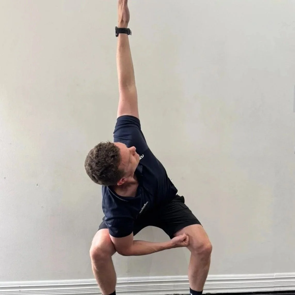 Man performing a yoga pose with one arm raised upright and the other arm bent with a clenched fist, standing against a plain wall.