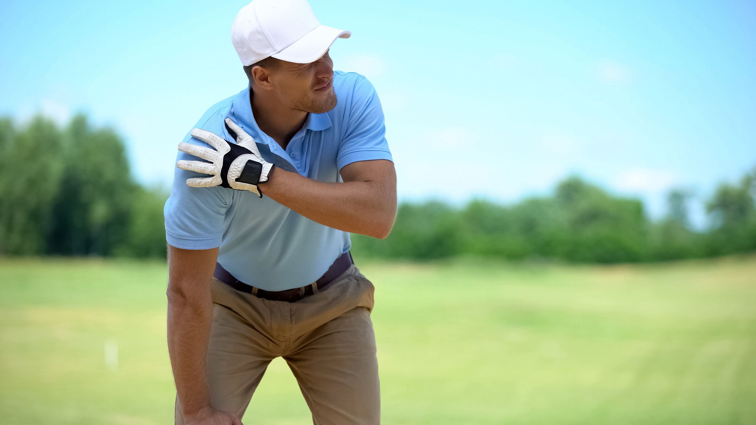 A man in a light blue polo shirt, khaki pants, and a white cap bending over on a golf course, resting his right hand on his shoulder while looking to his right. He is wearing a glove on his left hand, and the background is a green, open field with trees under a bright blue sky.