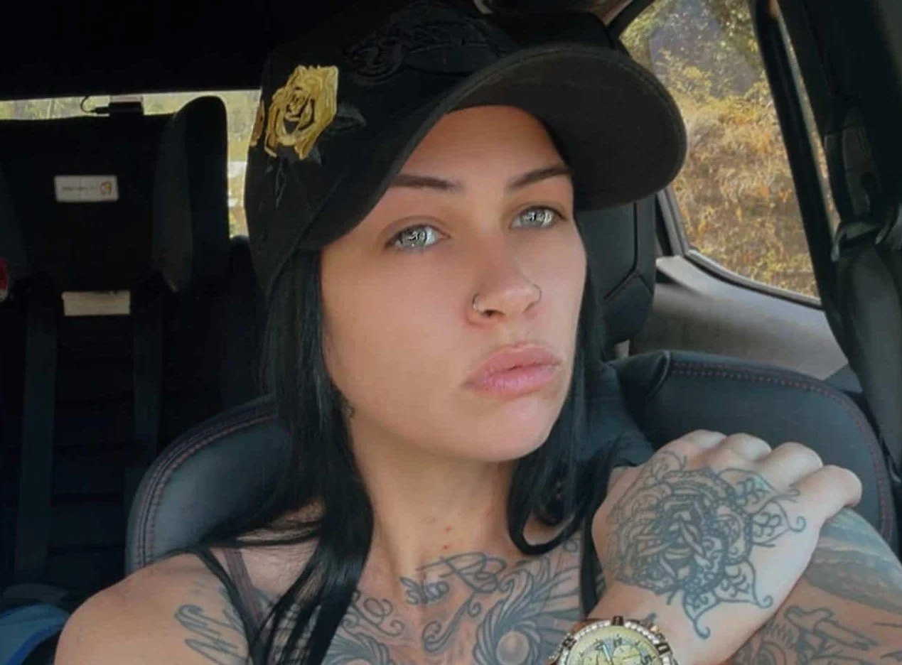 Woman with tattoos, wearing a black cap with a lion emblem and a gold watch, sitting in a vehicle with trees outside the window.