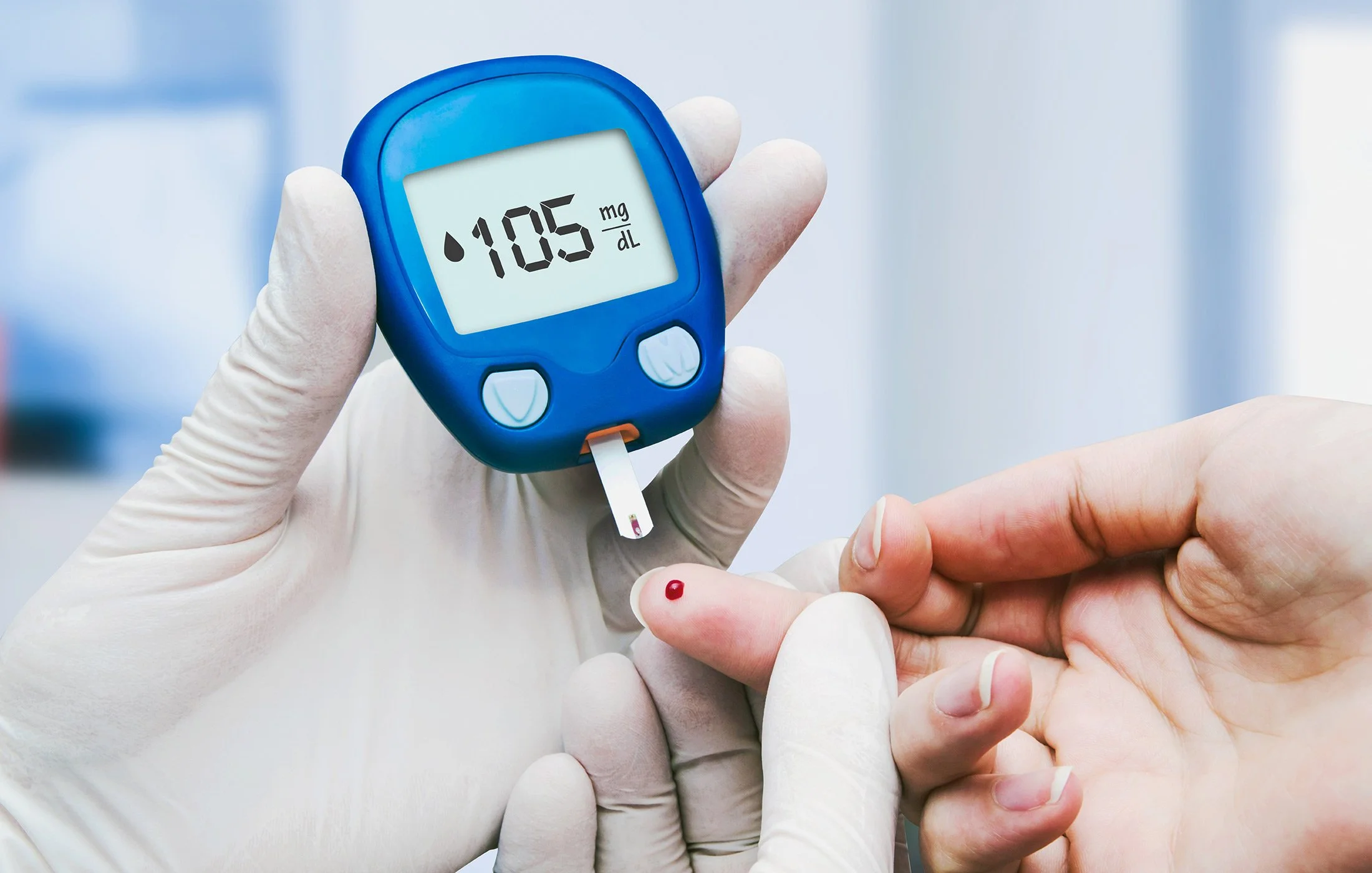 A healthcare professional wearing gloves uses a blood glucose meter to test blood sugar levels from a person's finger. The meter displays a reading of 105 mg/dL.