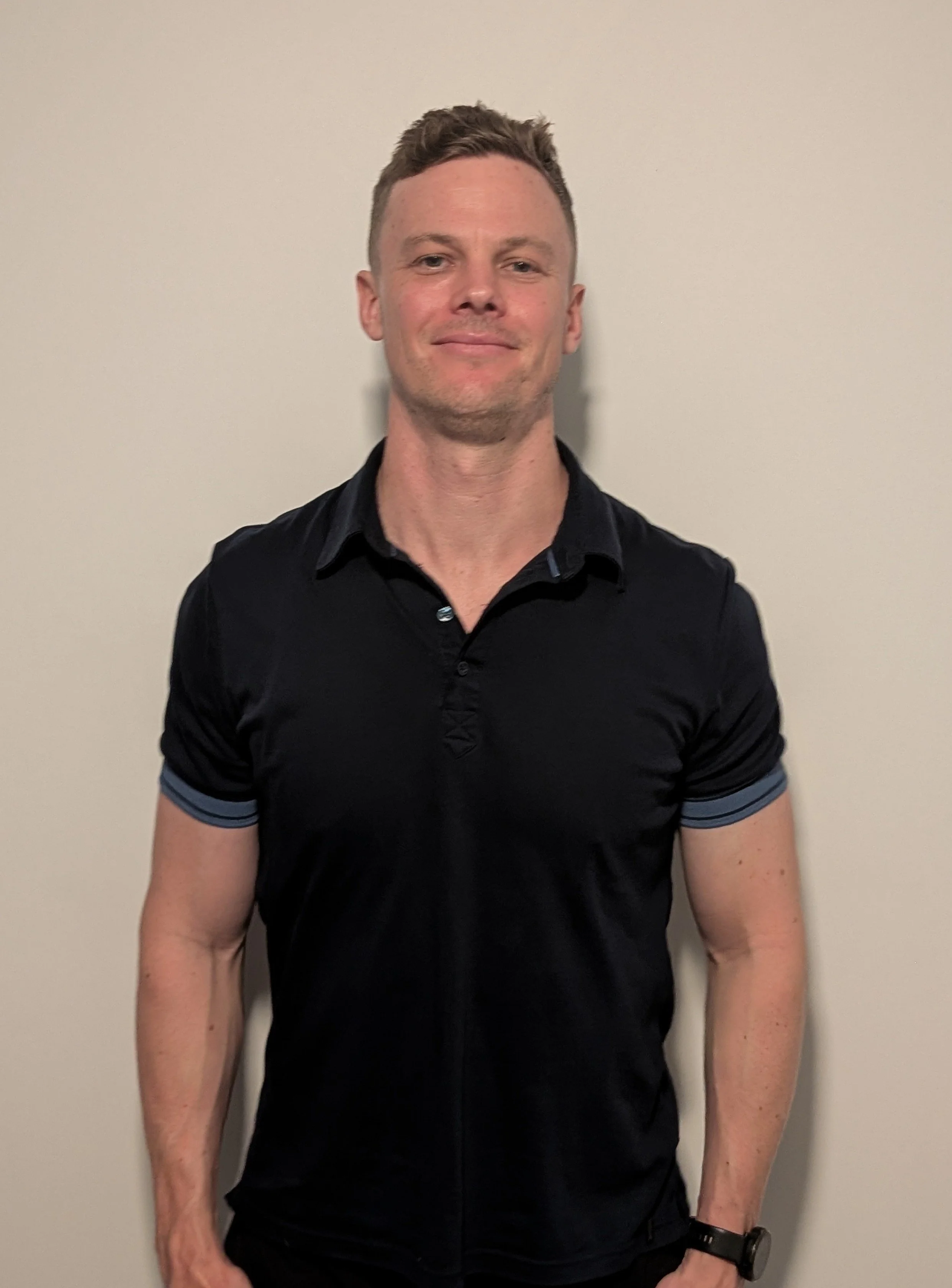 A man with short light brown hair, wearing a black polo shirt, smiling, standing against a plain light-colored wall.