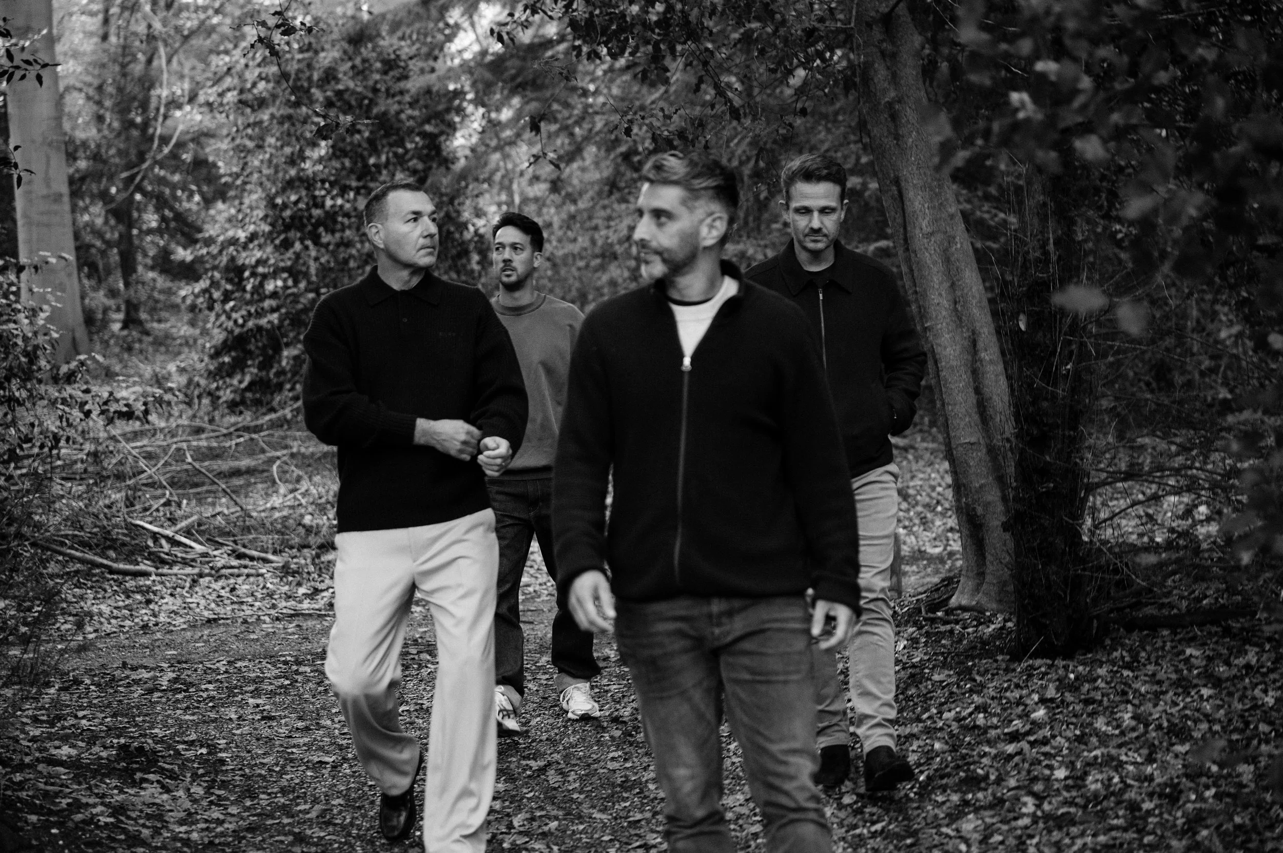 Four men walking through a wooded area on a trail, dressed casually, with trees and foliage around them in black and white.
