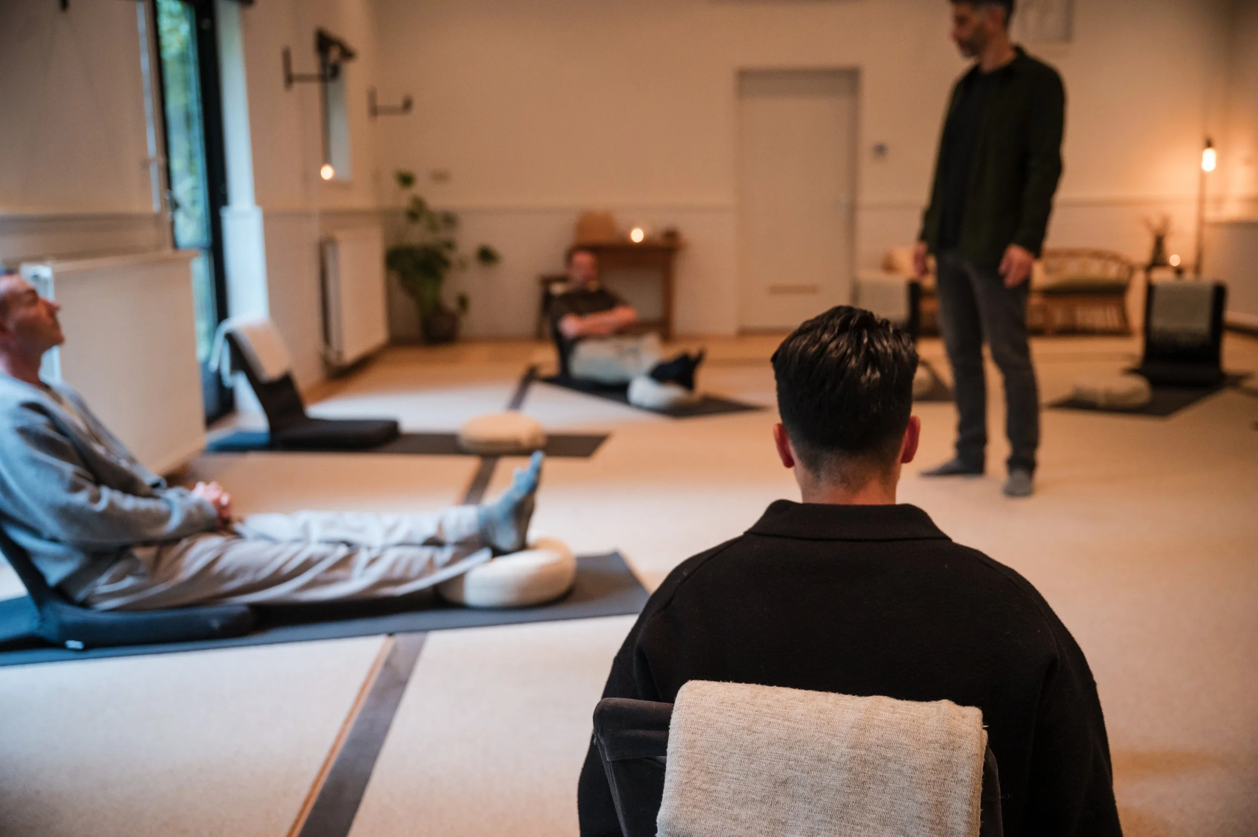 People practicing yoga or meditation in a spacious, softly lit room with mats, cushions, and gentle lighting, some sitting or lying on mats.