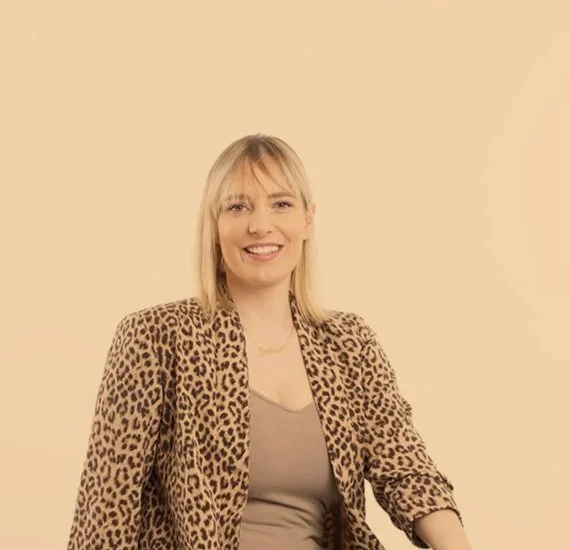 Eine junge Frau lächelt, trägt eine Leopardenmusterjacke und einen grauen Top, sitzt vor einem cremefarbenen Hintergrund.