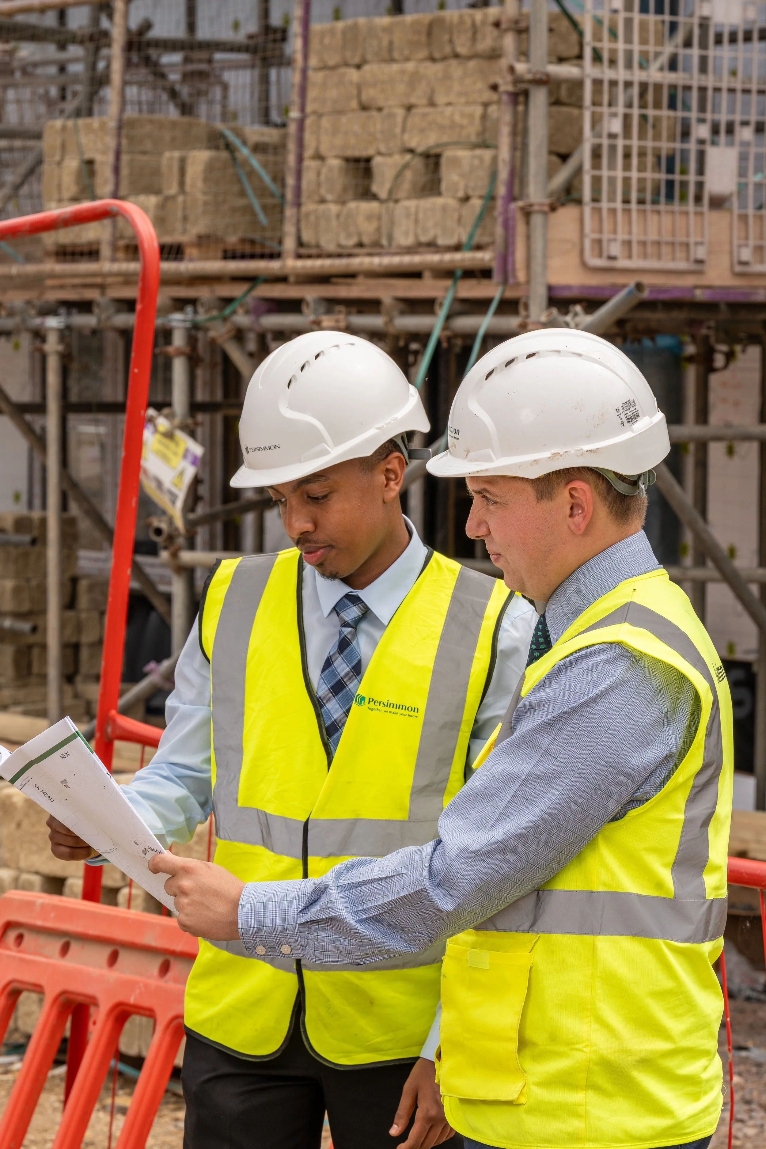 Two male construction workers wearing white safety helmets and yellow reflective vests discuss a blueprint at a construction site with scaffolding and building materials in the background.