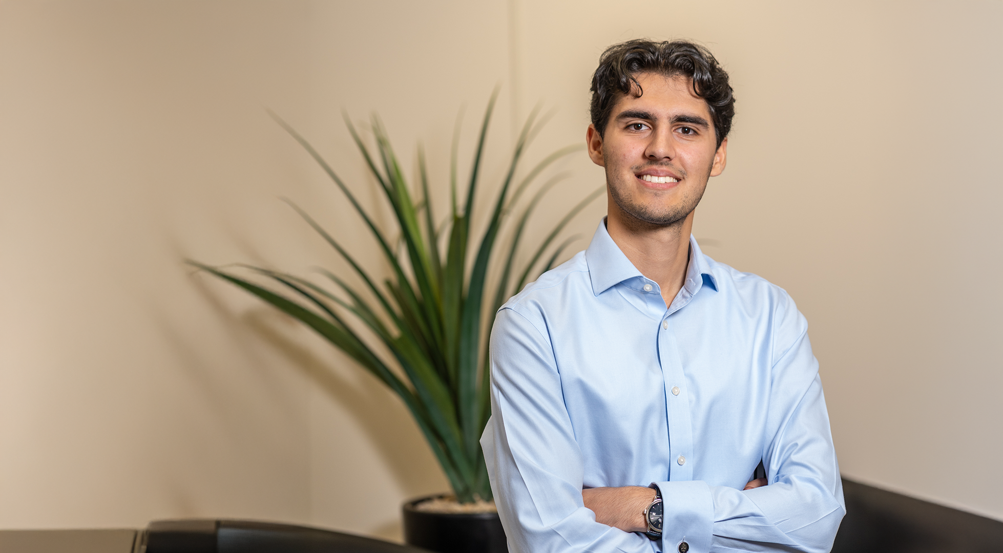 A young man with dark, curly hair, wearing a light blue dress shirt, standing with arms crossed in front of a potted plant with long green leaves, on a beige wall background.