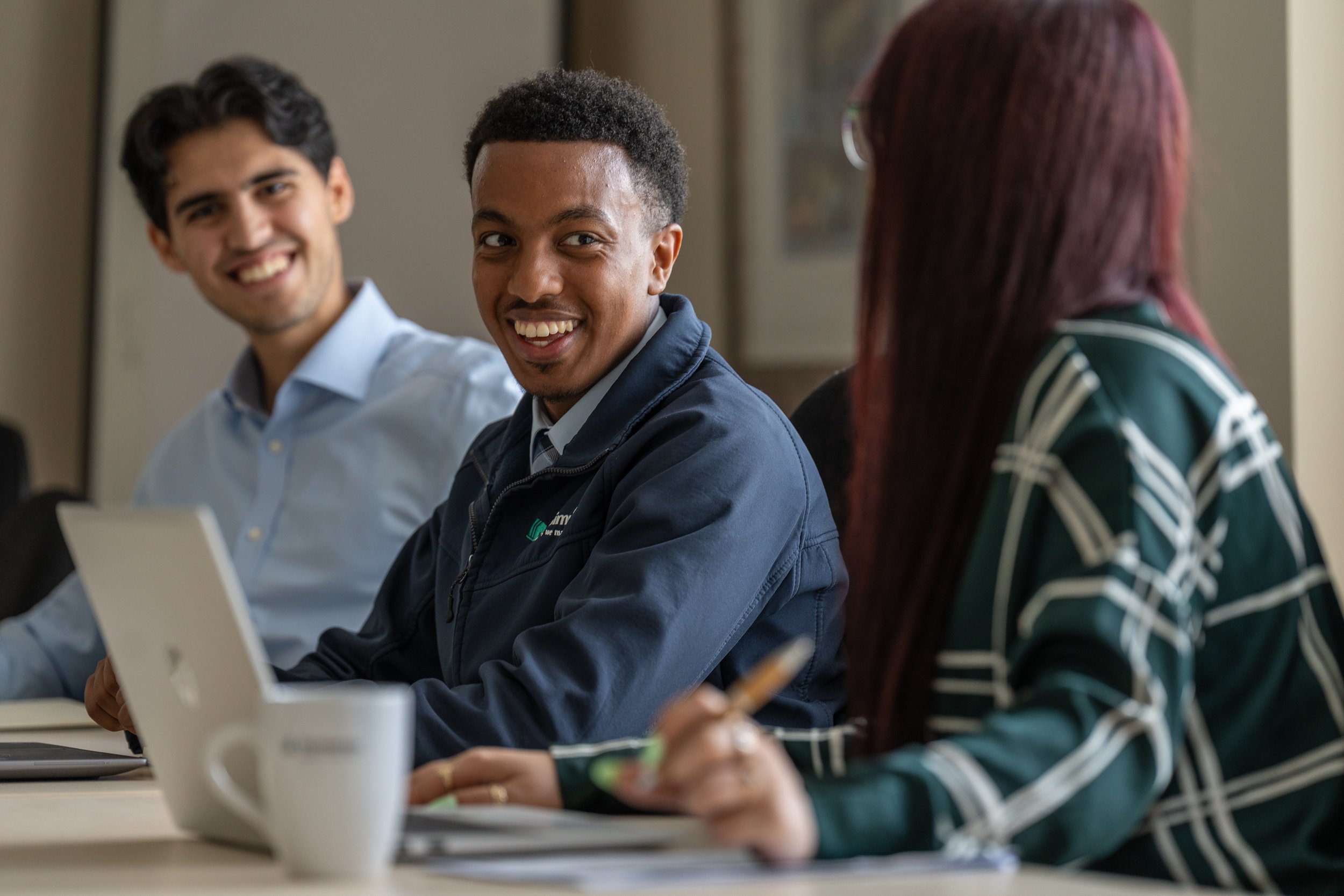 Three young adults sitting at a table during a discussion or meeting, smiling and engaging, with laptops, notebooks, and a mug visible on the table.