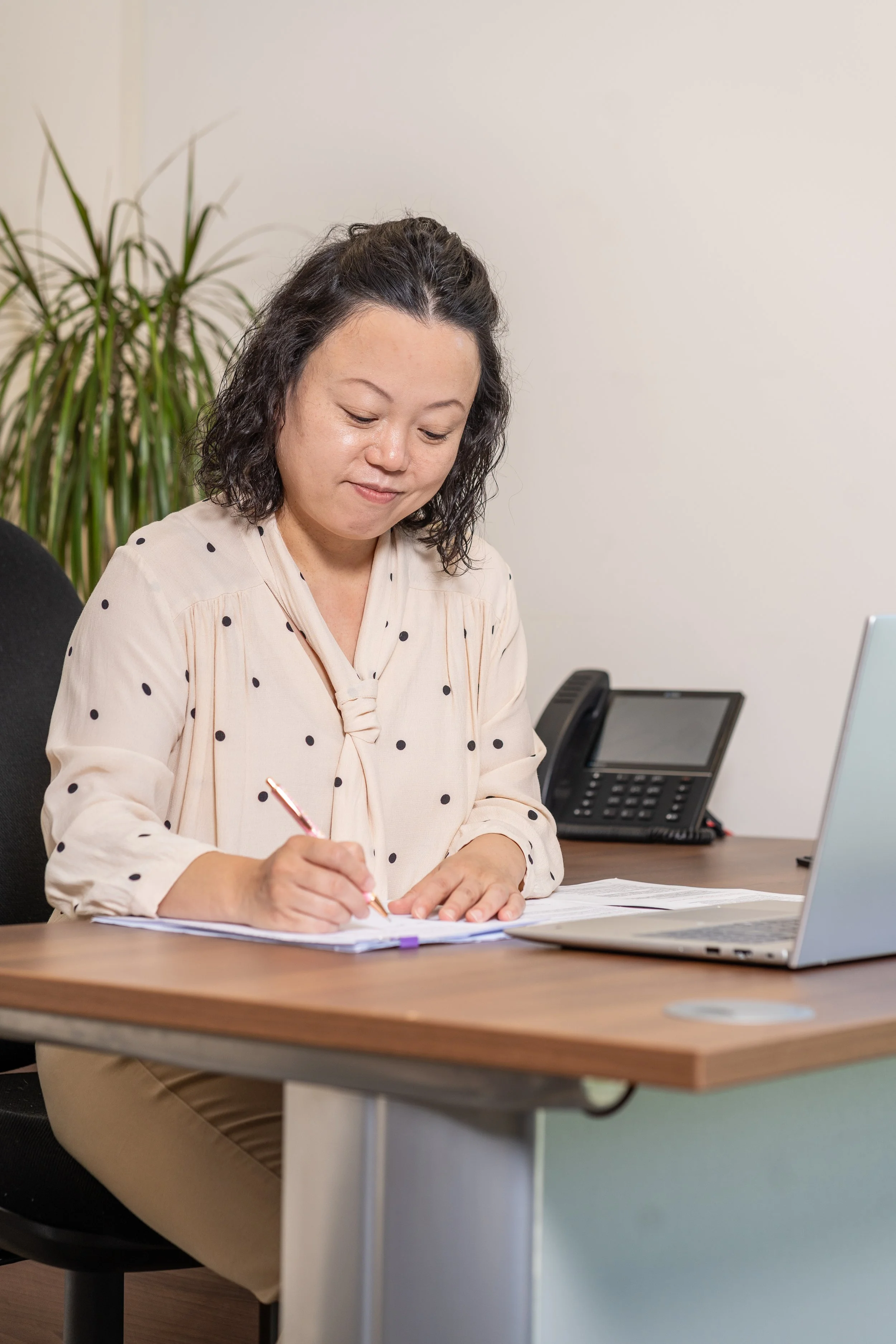 A woman sitting at a desk writing on a notepad with a pen, with a laptop and a phone on the desk, in an office with a plant in the background.