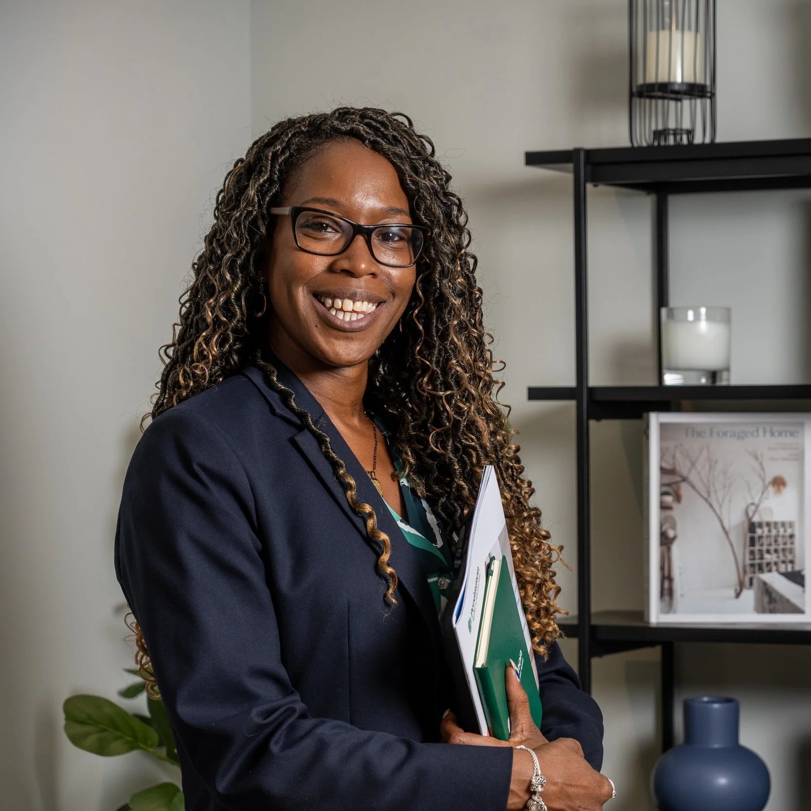 A woman with glasses, long curly hair, dressed in a dark blazer, holding notebooks, standing in an office or living room with bookshelf and decorative items.