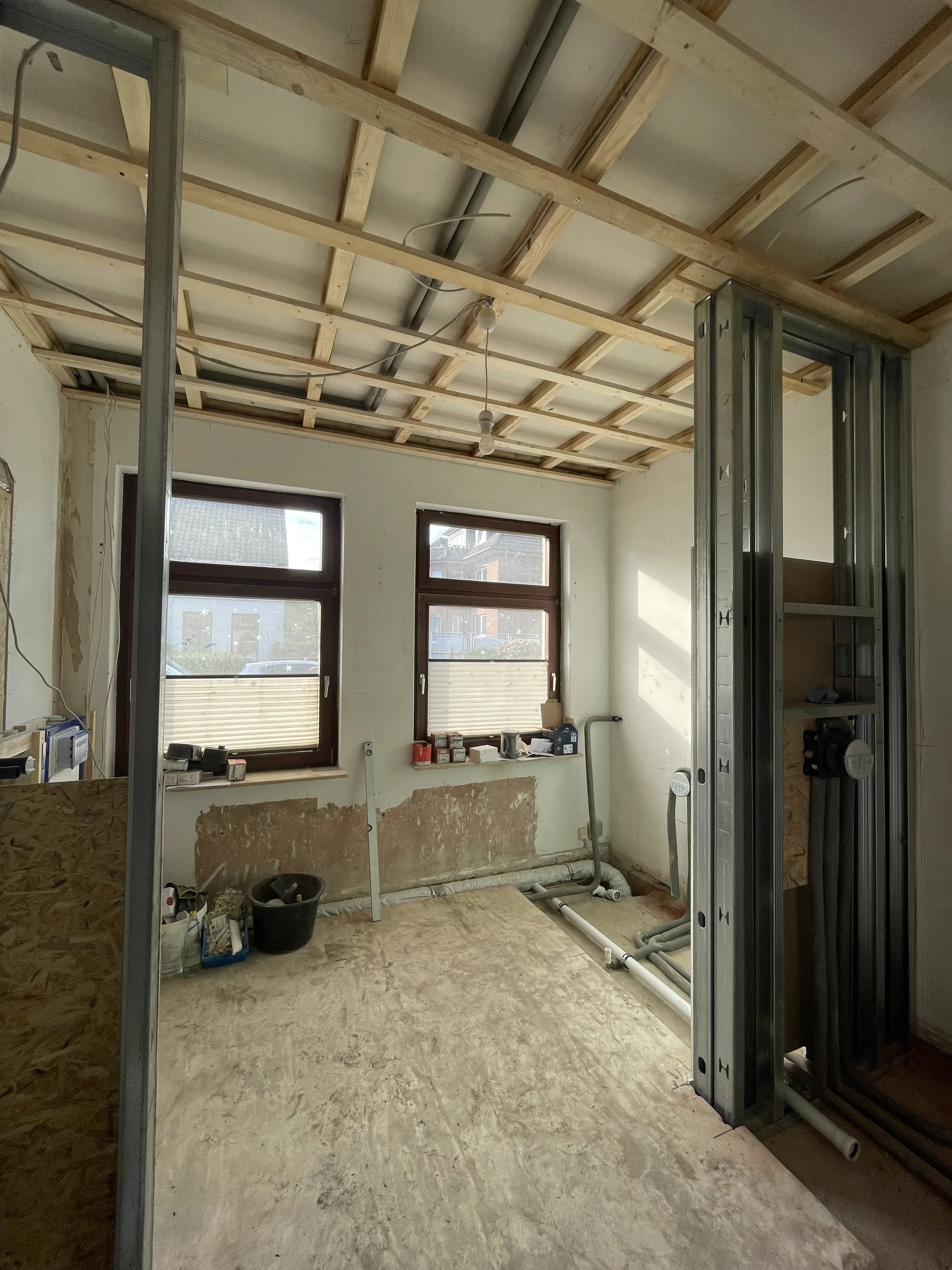 Trockenbau- und Rohinstallationsarbeiten bei einer Altbausanierung in Oldenburg. Zu sehen sind Metallprofile als Raumteiler, eine hölzerne Deckenlattung sowie erste Abwasserrohre und Baumaterialien vor zwei Fenstern.