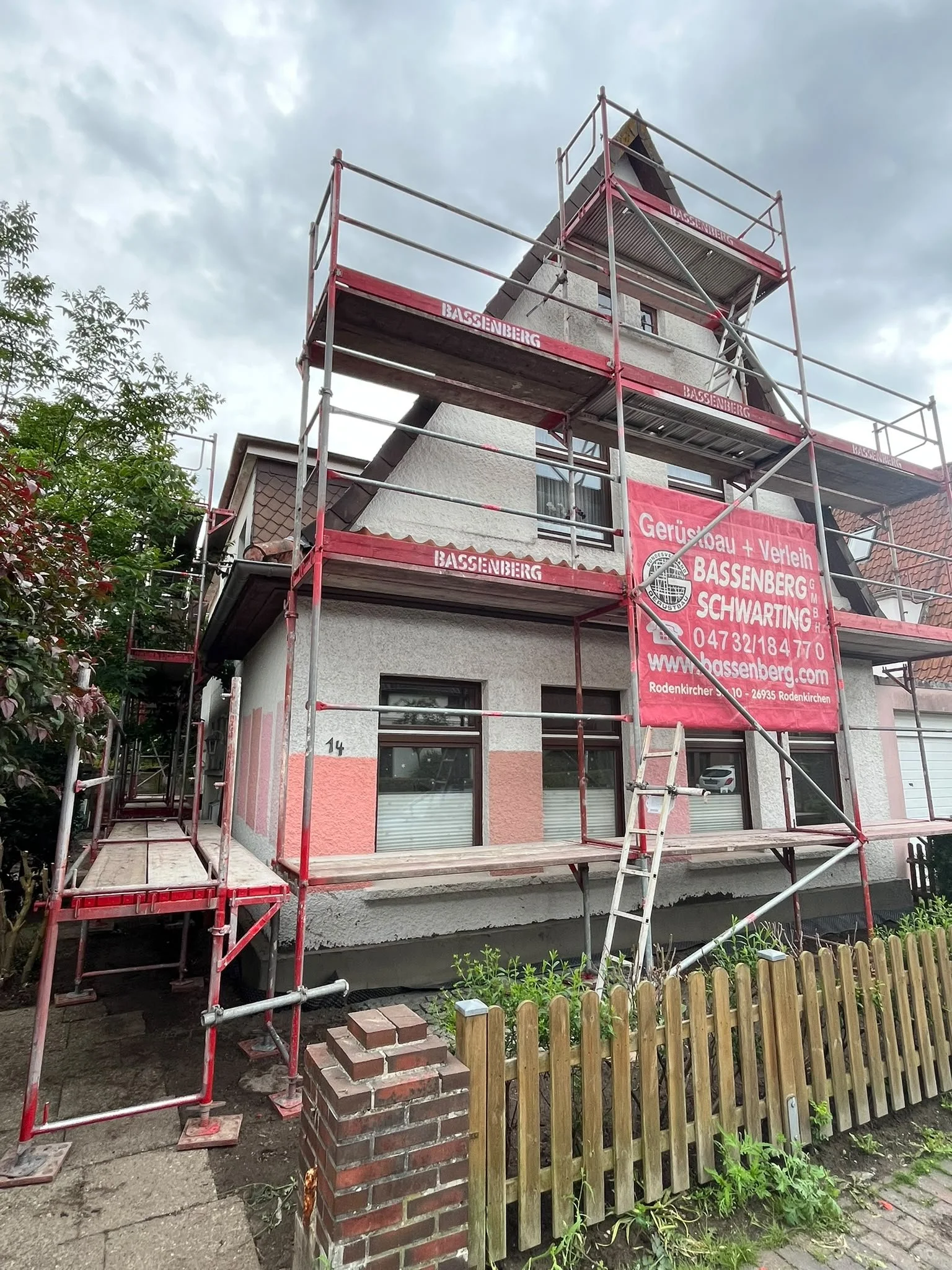 Altbausanierung in Oldenburg: Ein komplett eingerüstetes Einfamilienhaus. An der Fassade steht ein rotes Baugerüst mit einem Banner der Firma Bassenberg. Im Vordergrund grenzt ein klassischer Holzzaun mit gemauerten Pfeilern die Baustelle ab.