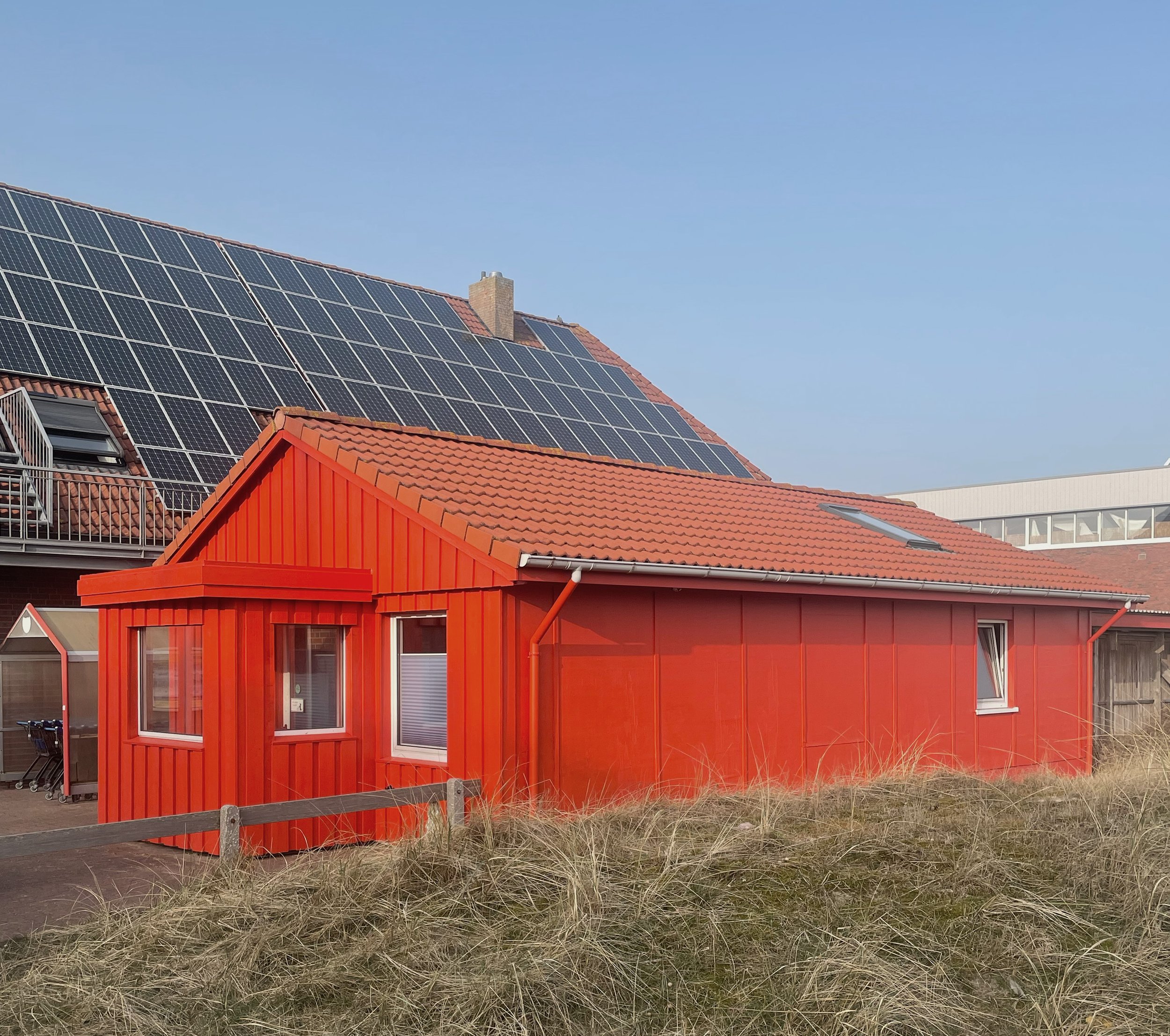 Kontrastreiche Architektur: Ein traditionelles, leuchtend rotes Holzgebäude steht vor einem modernen Dach mit flächendeckenden Solarpanels, umgeben von Küstengras.