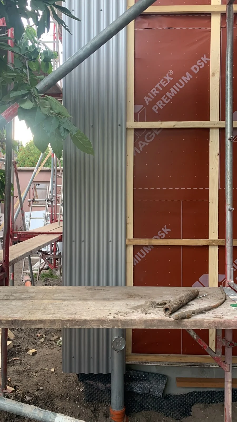 Montage einer vorgehängten Fassade bei einer Altbausanierung in Oldenburg. Das Bild zeigt den Übergang von der roten Winddichtbahn mit Konterlattung zur fertig verschraubten grauen Wellblechverkleidung, fotografiert von einem Baugerüst aus.