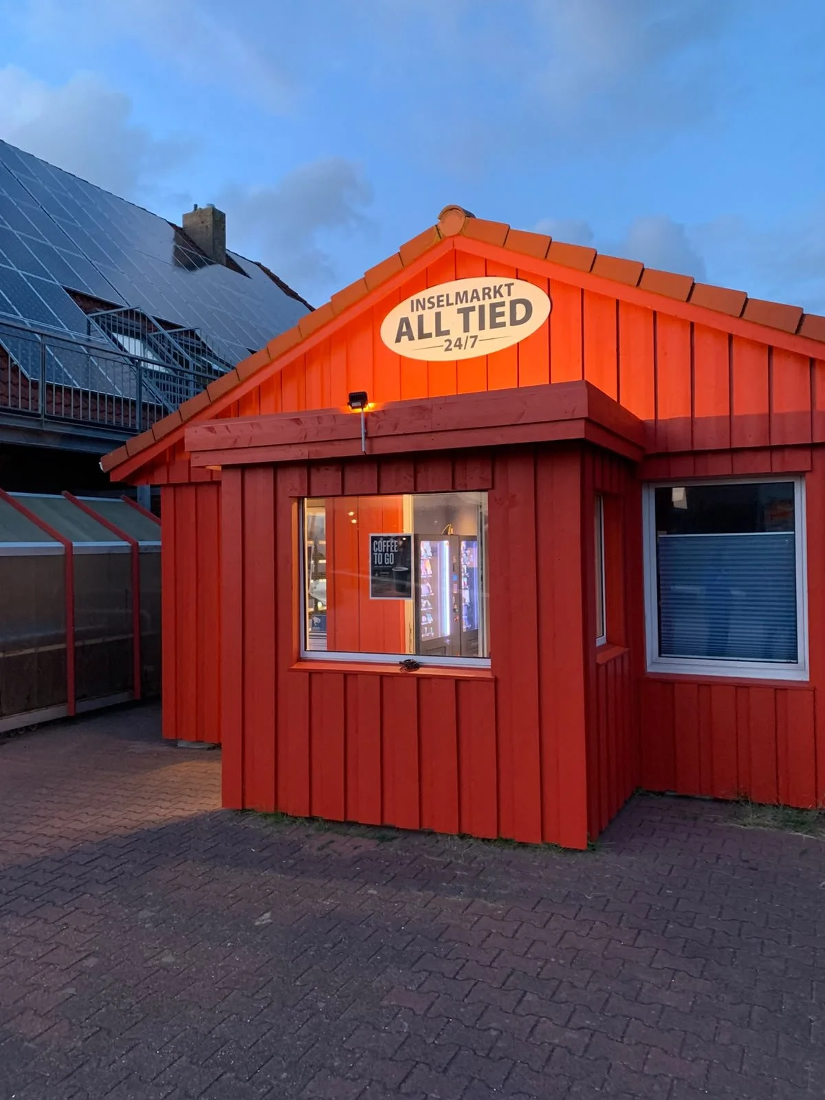 Die fertig umgebaute ehemalige Backstube auf Baltrum: Das Gebäude erstrahlt nun als roter 24/7-Inselmarkt namens 'All Tied'. Die abendliche Beleuchtung hebt das Schild und das Schaufenster mit den beleuchteten Snack- und Kaffeeautomaten hervor.