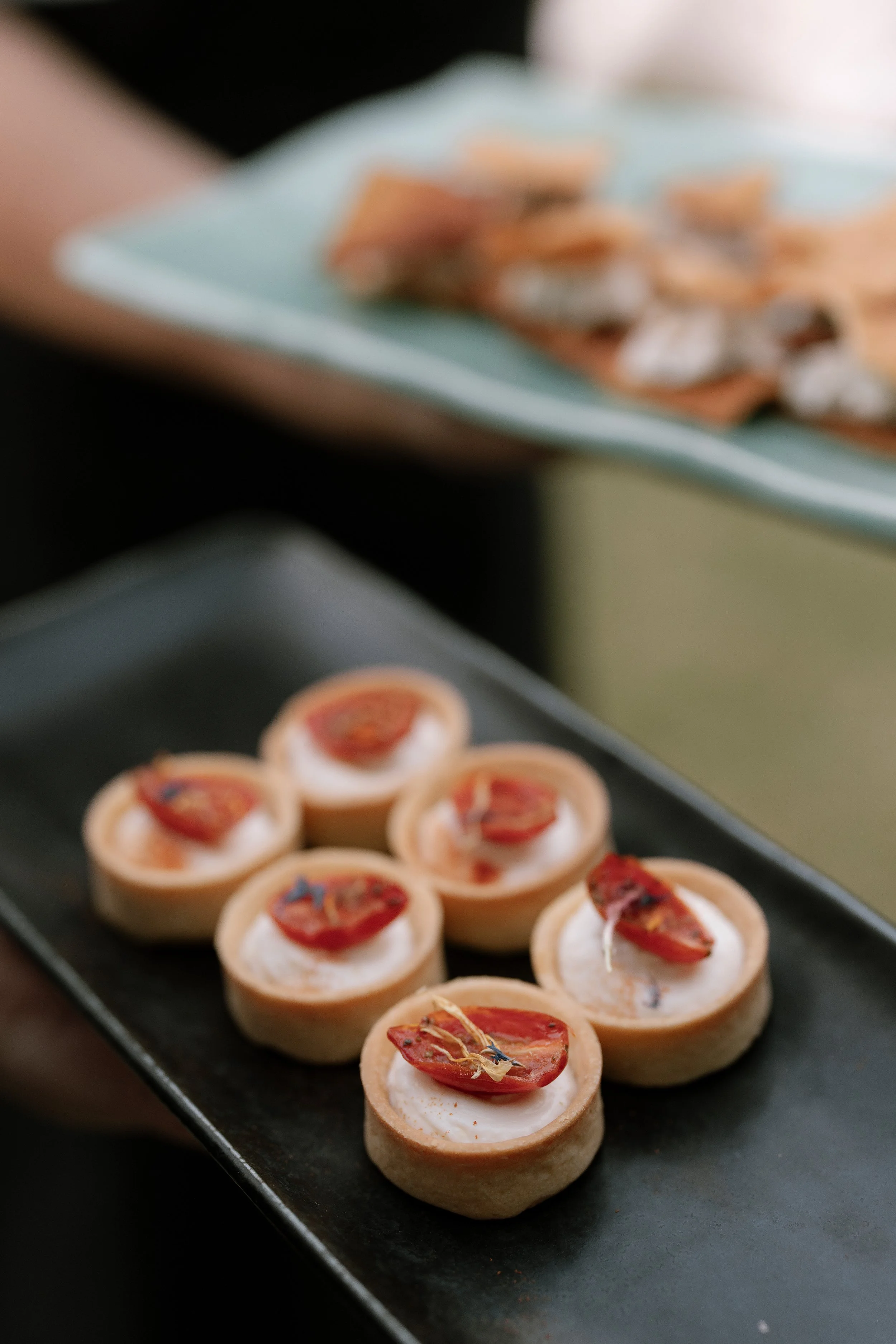 Miniature savory tarts topped with cherry tomato slices on a black serving platter.