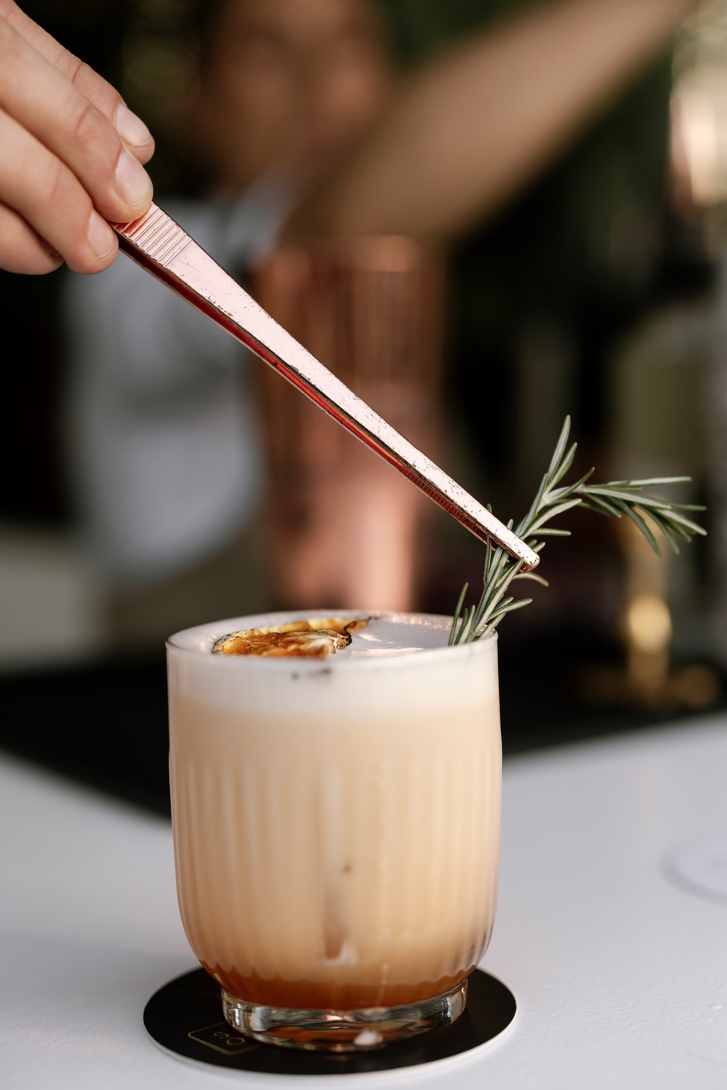 A person holds a metallic tool, likely a bar spoon or stirring rod, garnishing a creamy cocktail with a sprig of rosemary and a slice of dried fruit in a clear glass.