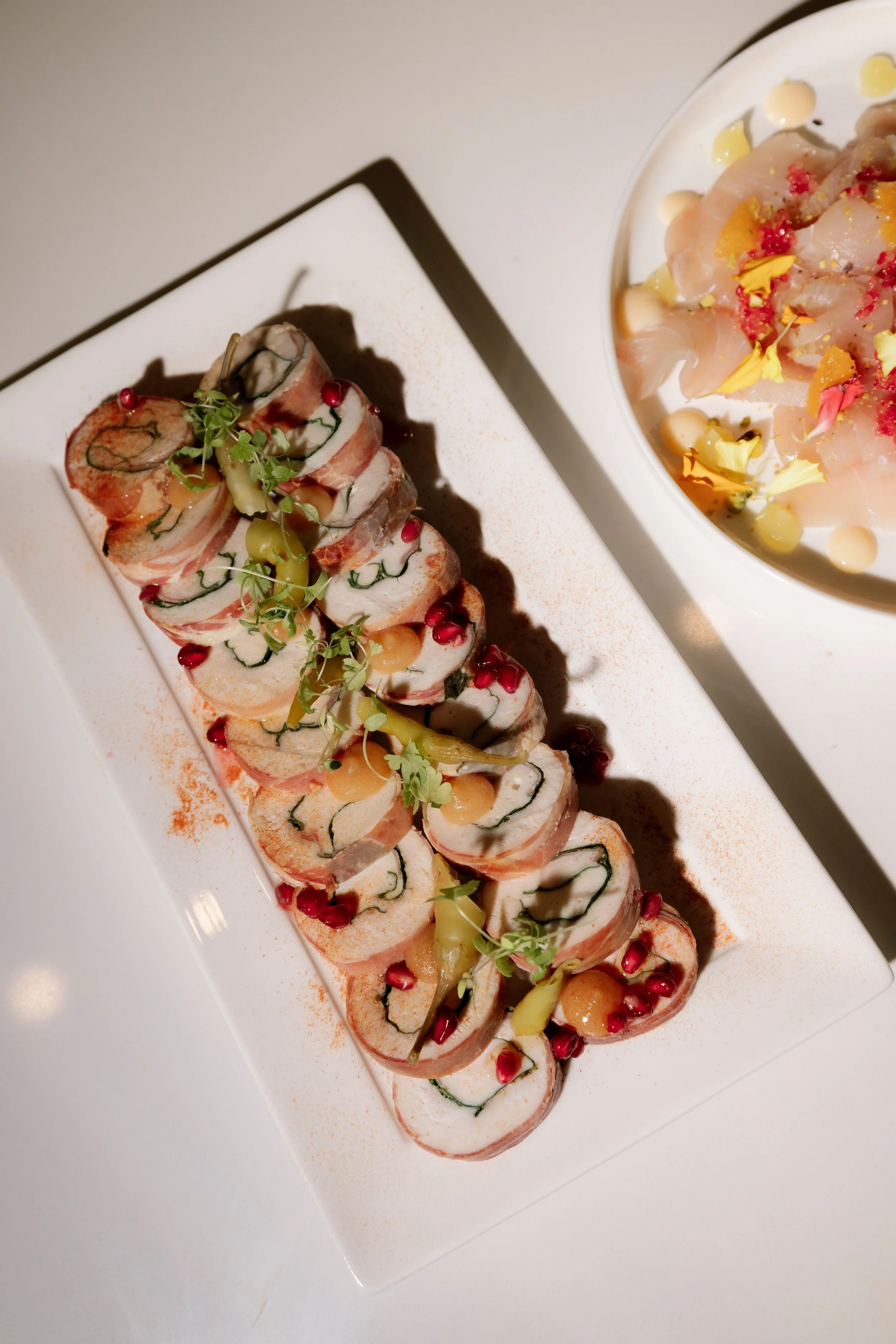 A rectangular white plate with sliced rolled appetizer garnished with microgreens, pomegranate seeds, and yellow pickled vegetables, next to a white bowl of sashimi with flower petals, nuts, and small sauce dots.