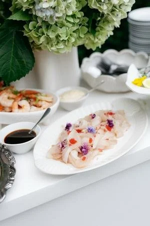 A platter of sashimi garnished with edible flowers, accompanied by soy sauce and wasabi, with a floral centerpiece and stacked white plates in the background.