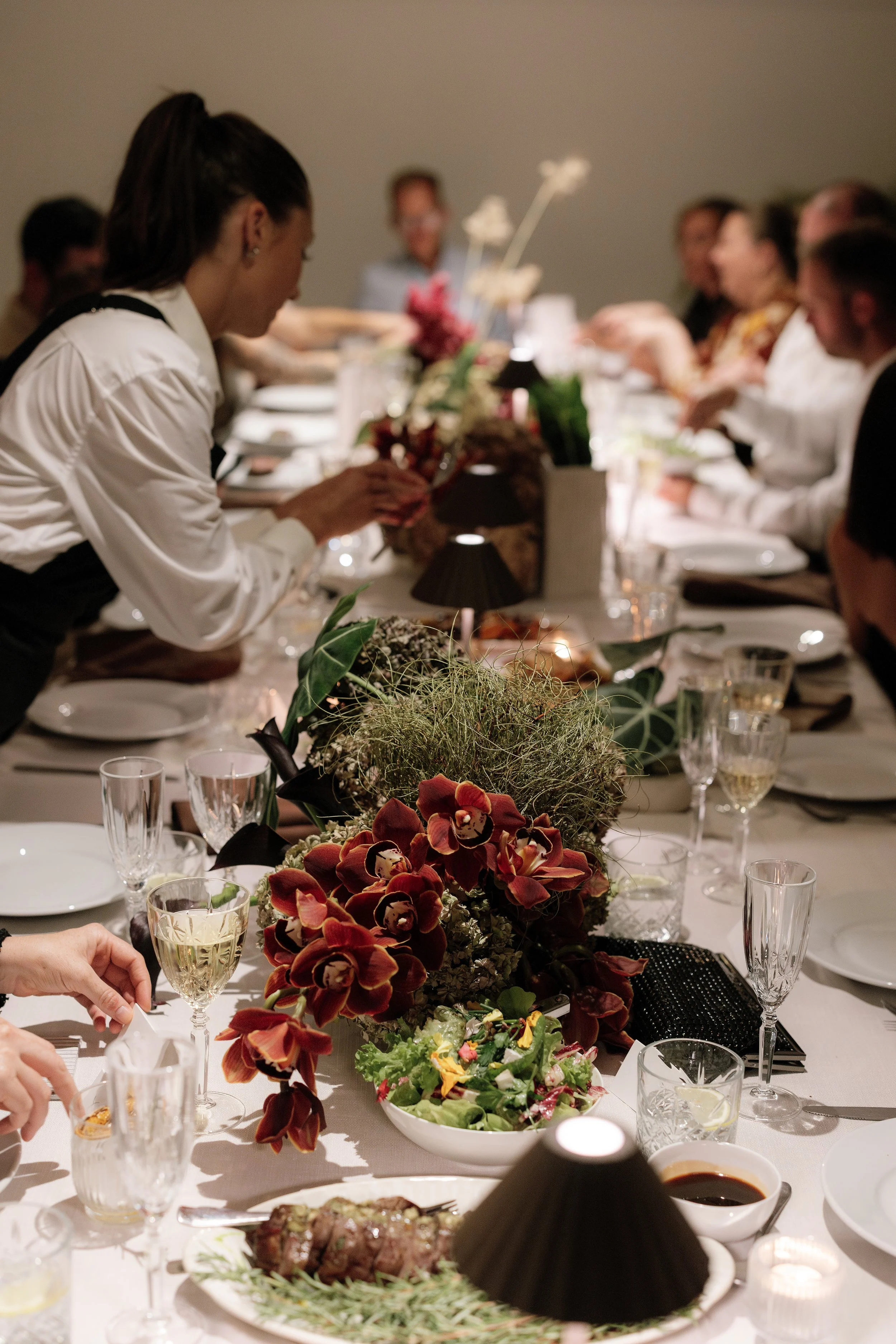 People seated at a long dinner table with floral centerpieces, glasses of wine, and plates of food, during a formal, yet warm and inviting gathering.