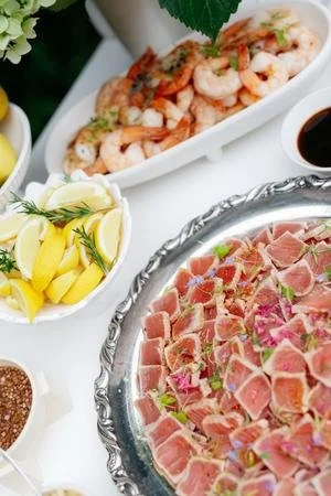 A plate of sliced prosciutto, a bowl of cooked shrimp, and a dish with lemon wedges, soy sauce, and a small bowl of dark sauce on a white table.