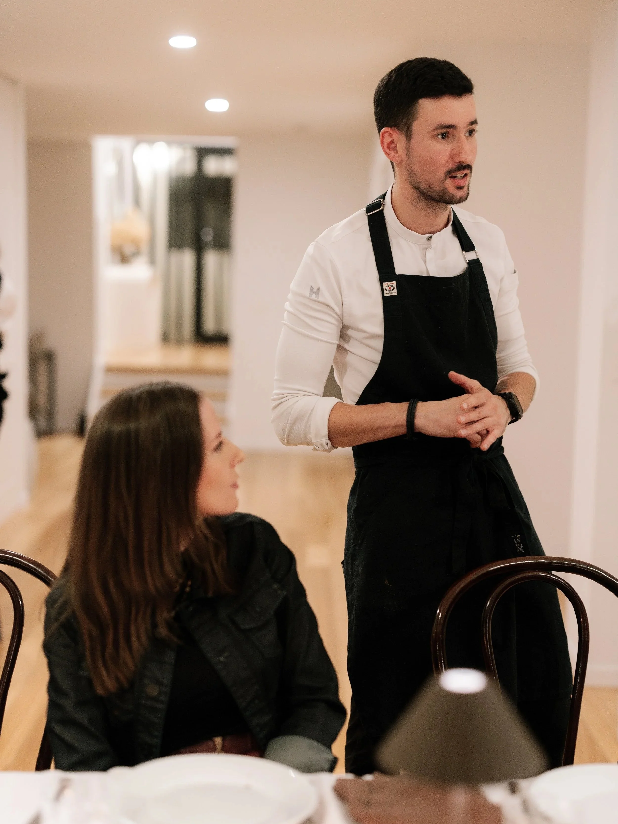 A Chef wearing a white shirt and black apron standing and explaining a menu to diners, with a woman sitting at a table ready to enjoy her meal.