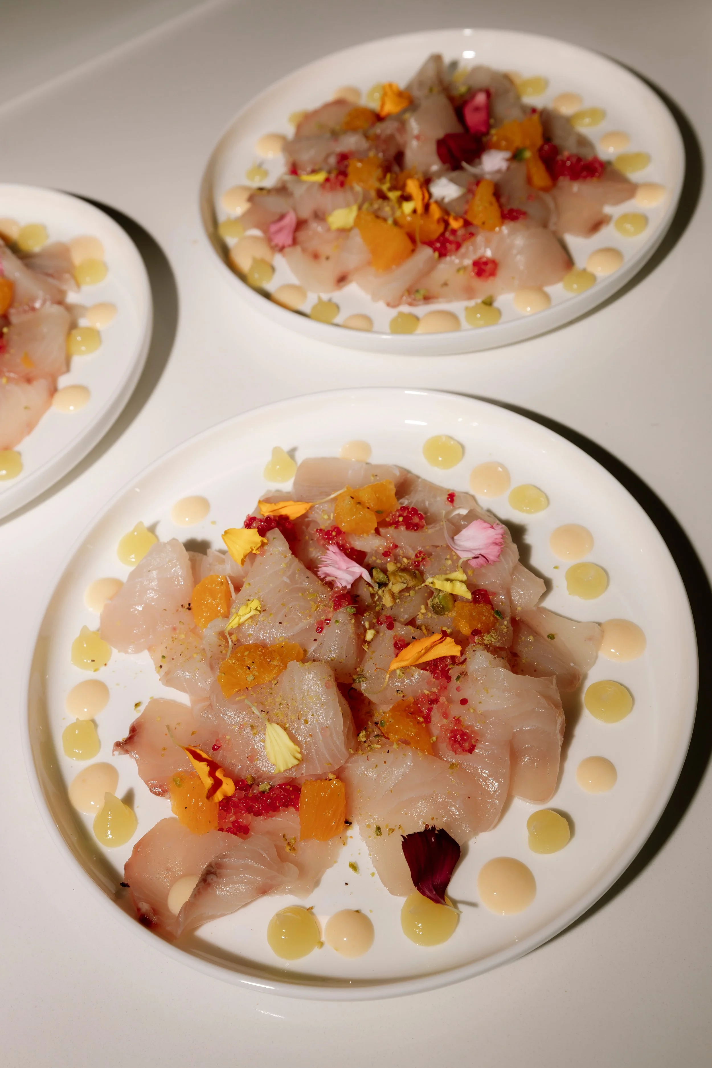 Plates of raw fish garnished with edible flowers, diced fruit, and dotted with sauce.