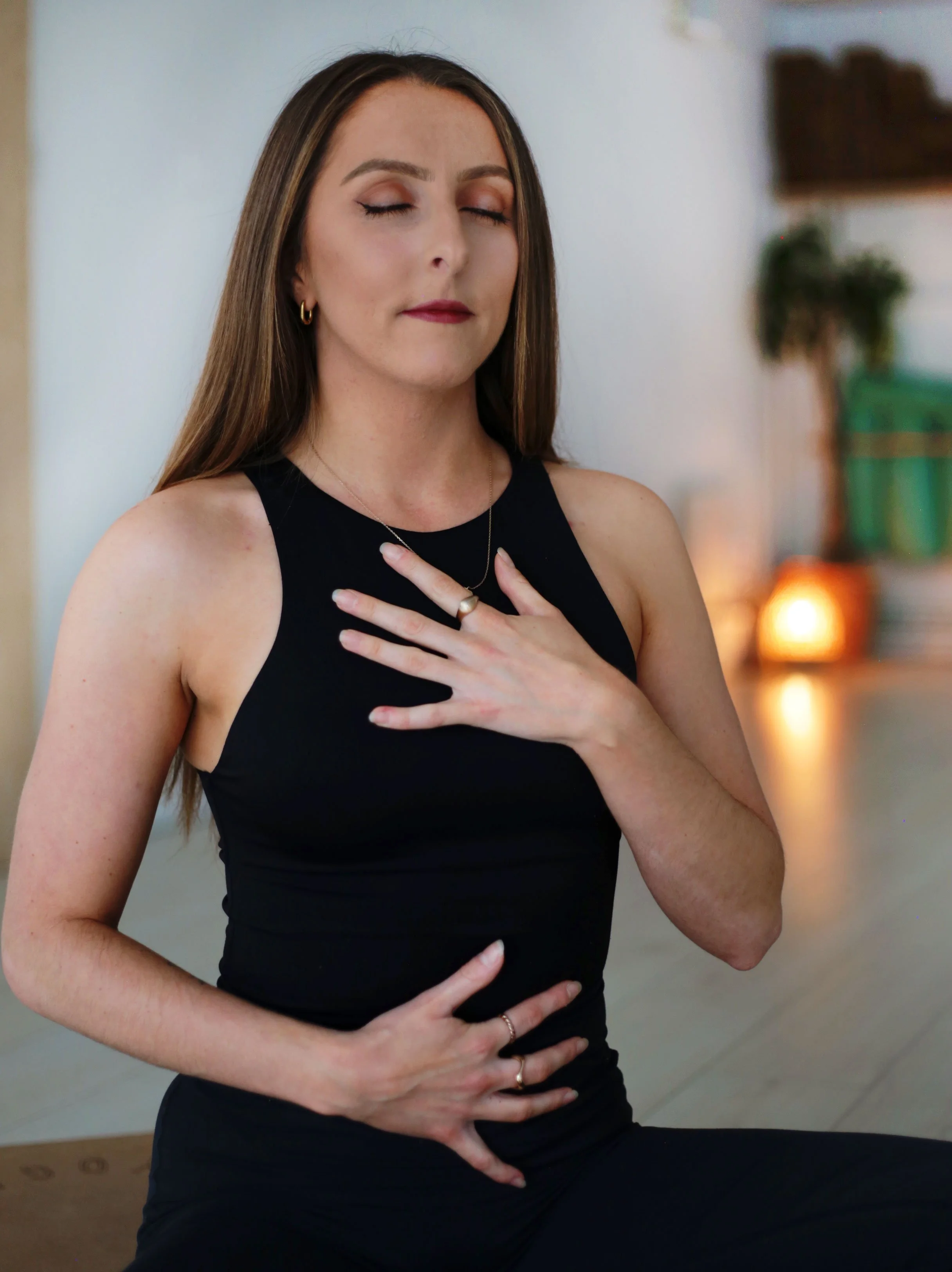 A woman practicing yoga in a side plank pose, wearing a black tank top and black pants, with long brown hair, an arm tattoo, and a neutral background.